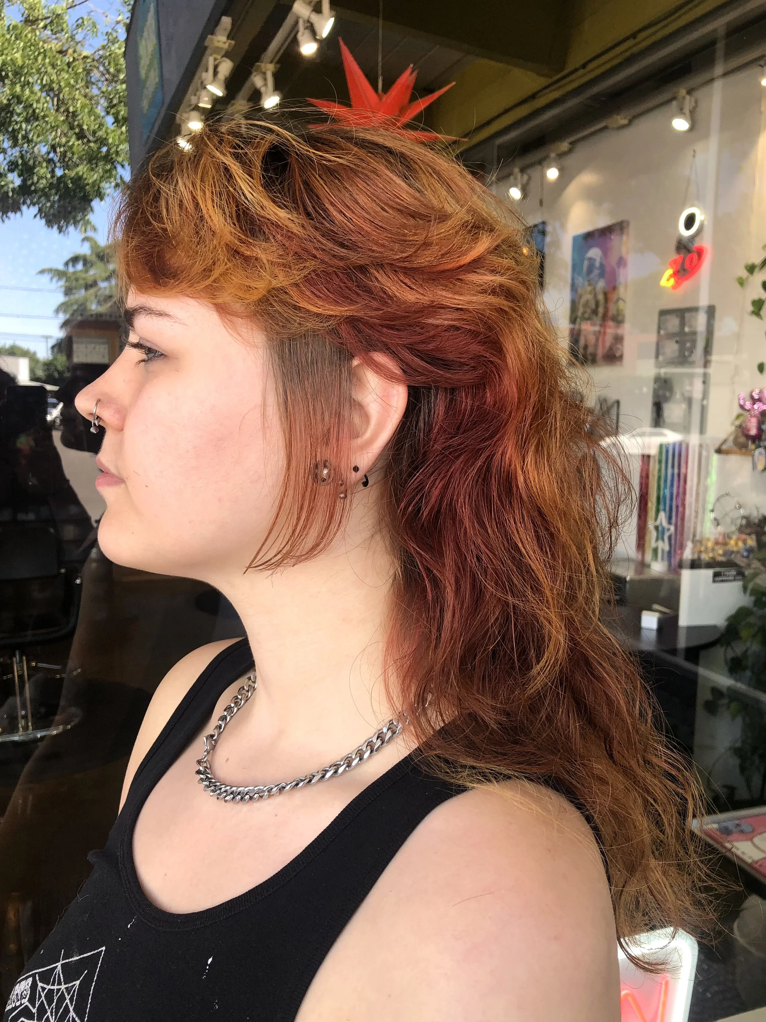 A woman with red and orange wavy hair is standing outside a storefront with a large glass window. She is wearing a black tank top, a silver chain necklace, and small earrings.