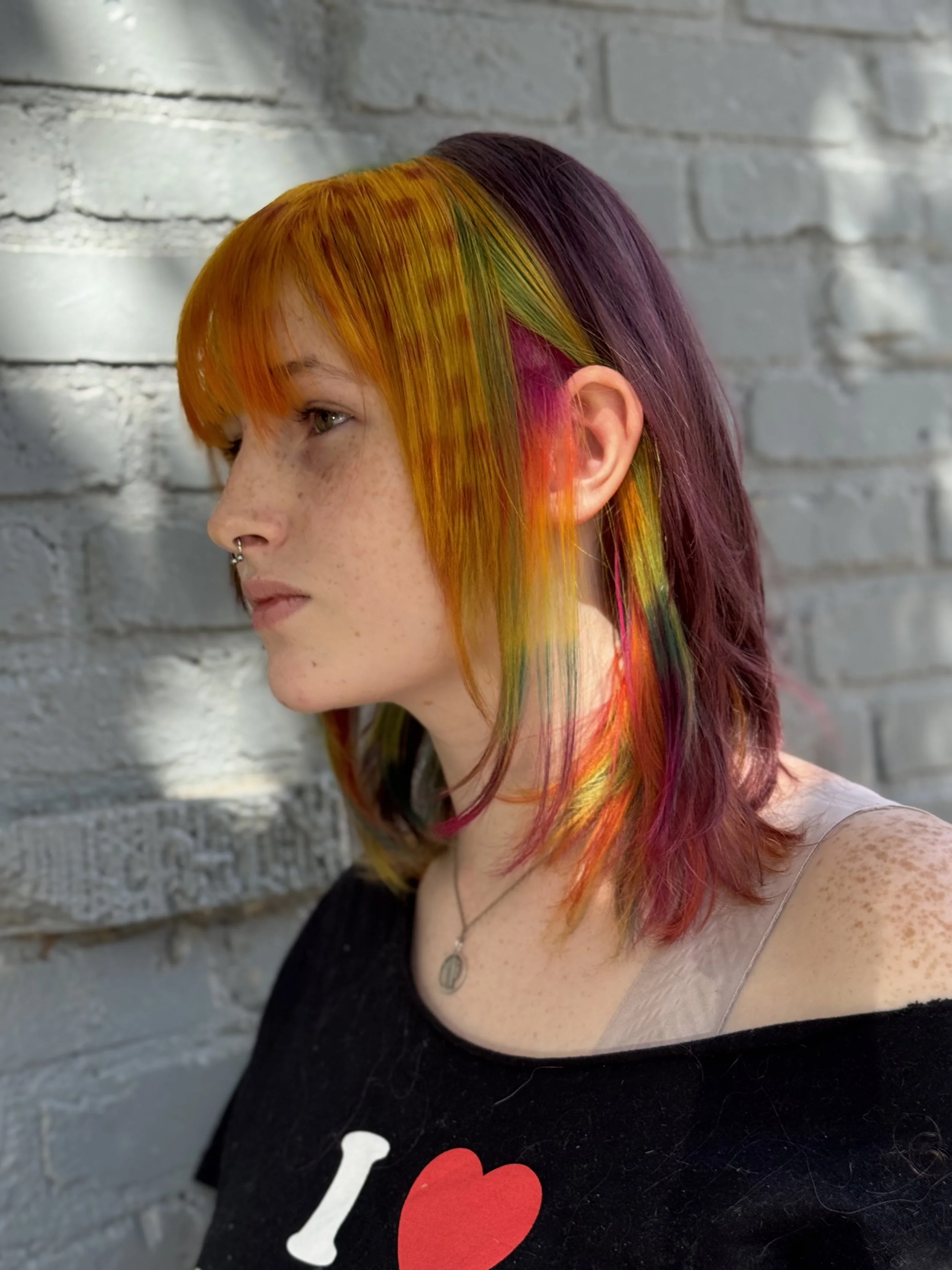 Side profile of a woman with multicolored hair in shades of orange, yellow, green, purple, and pink, standing against a gray brick wall.