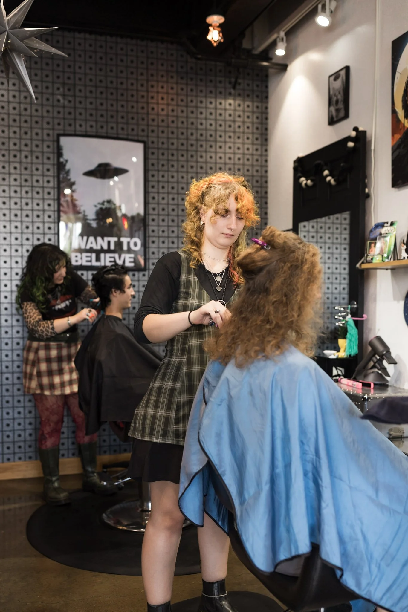 A young woman getting her hair styled by a hairstylist in a salon. The salon has patterned wallpaper, framed pictures, and modern lighting.