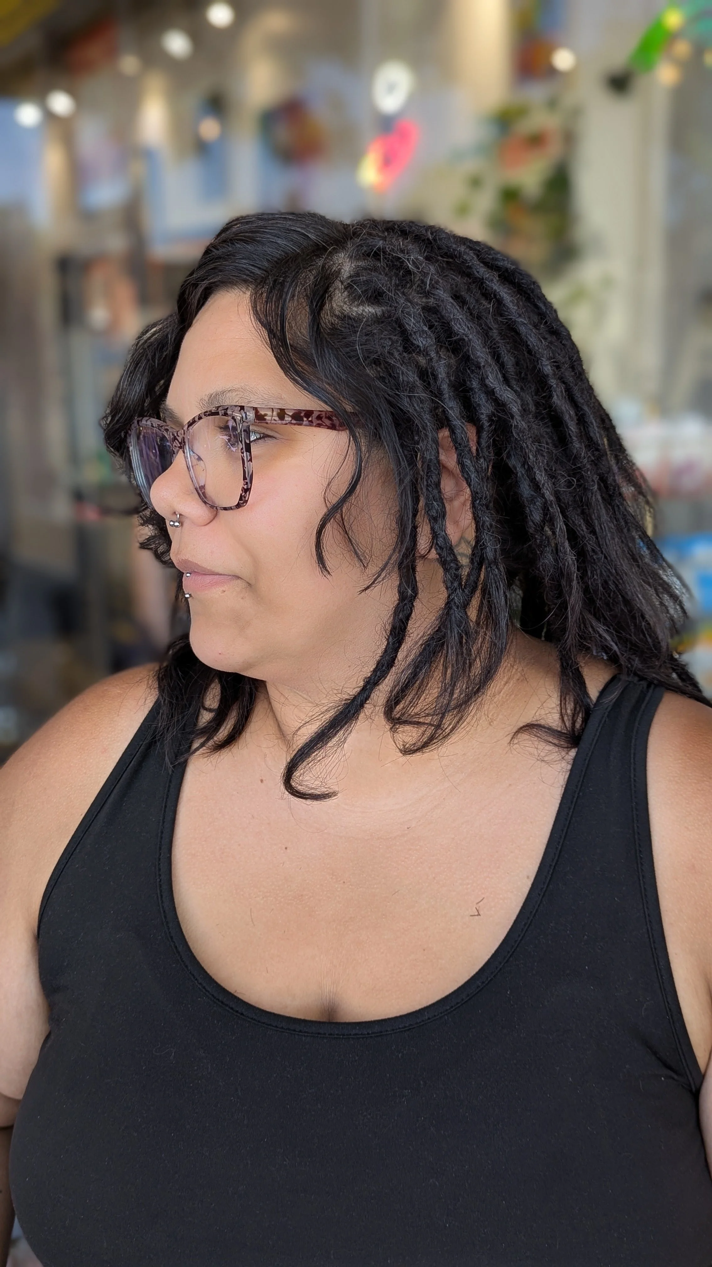 Side profile of a woman with black hair styled into dreadlocks, wearing glasses and a black tank top, in an indoor setting with blurred background.