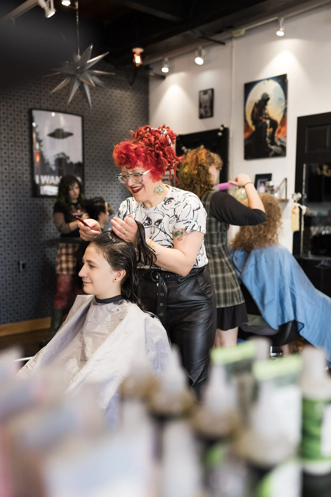 A hairdresser with red and orange curly hair, wearing glasses and earrings, styling a young woman's dark hair at a salon, with other clients and hairdressers in the background.