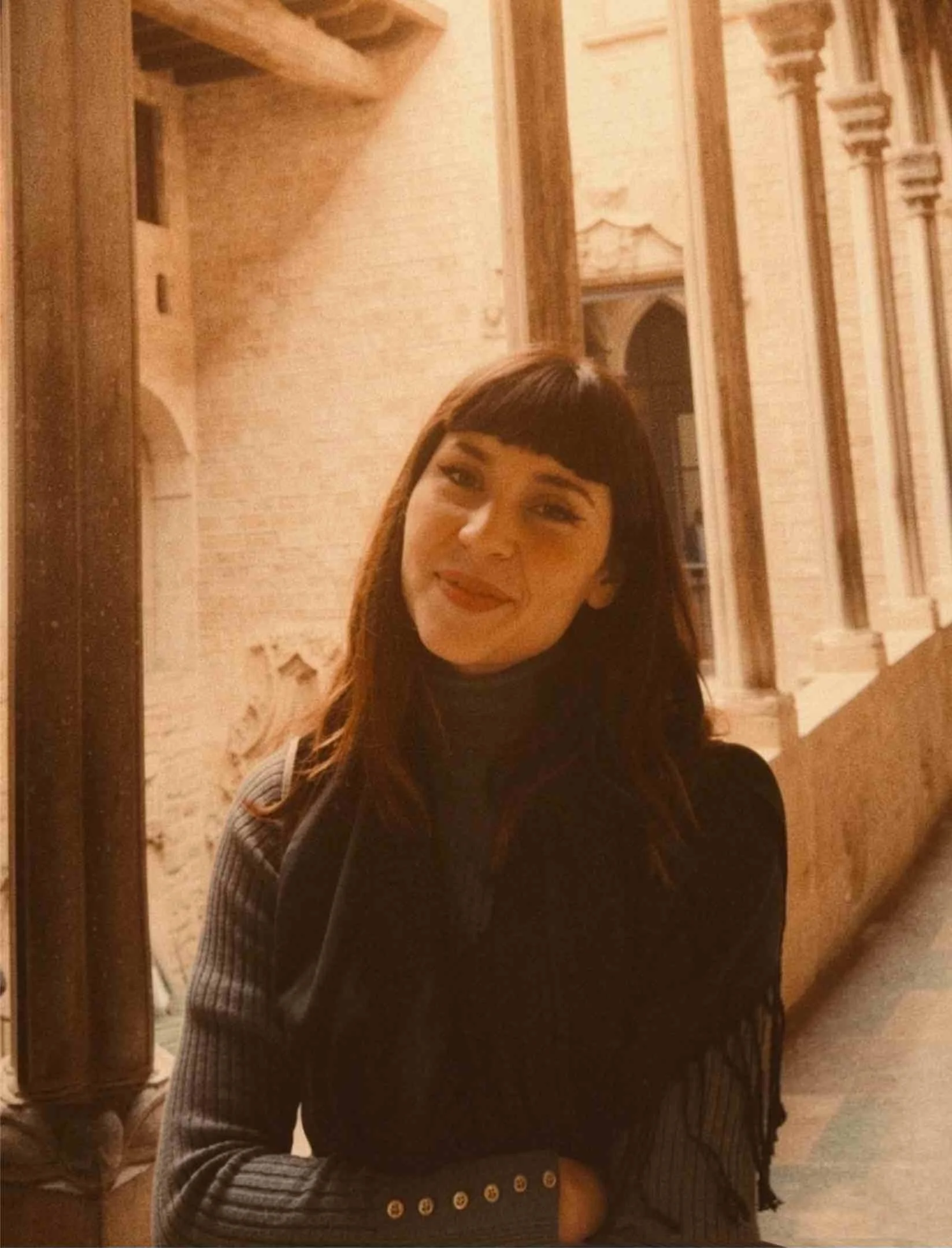 A woman wearing a black turtleneck with shoulder length brown hair smiles, her head tilted to the right, in front of historic white building.