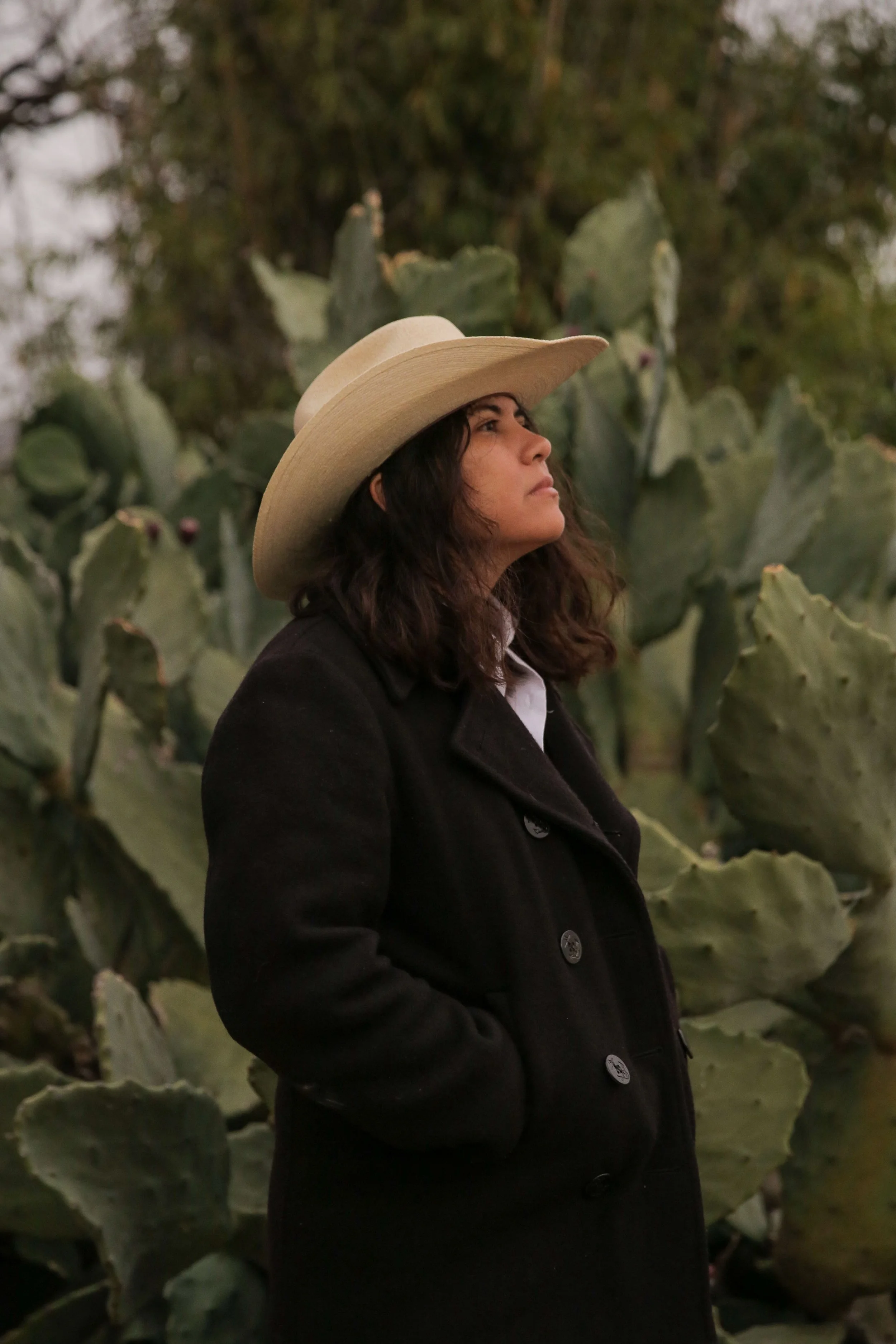 A woman with shoulder length brown and wavy hair wearing a light colored cowboy hat, white shirt, and black jacket,  stands in profile and is looking out in front of a field of nopales.