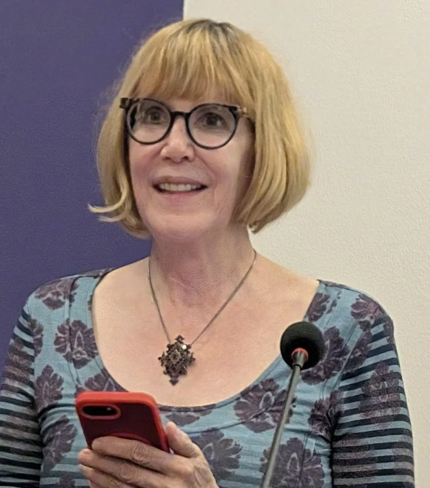 woman wearing glasses and large pendant necklace  looking slightly upward from behind podium with mic