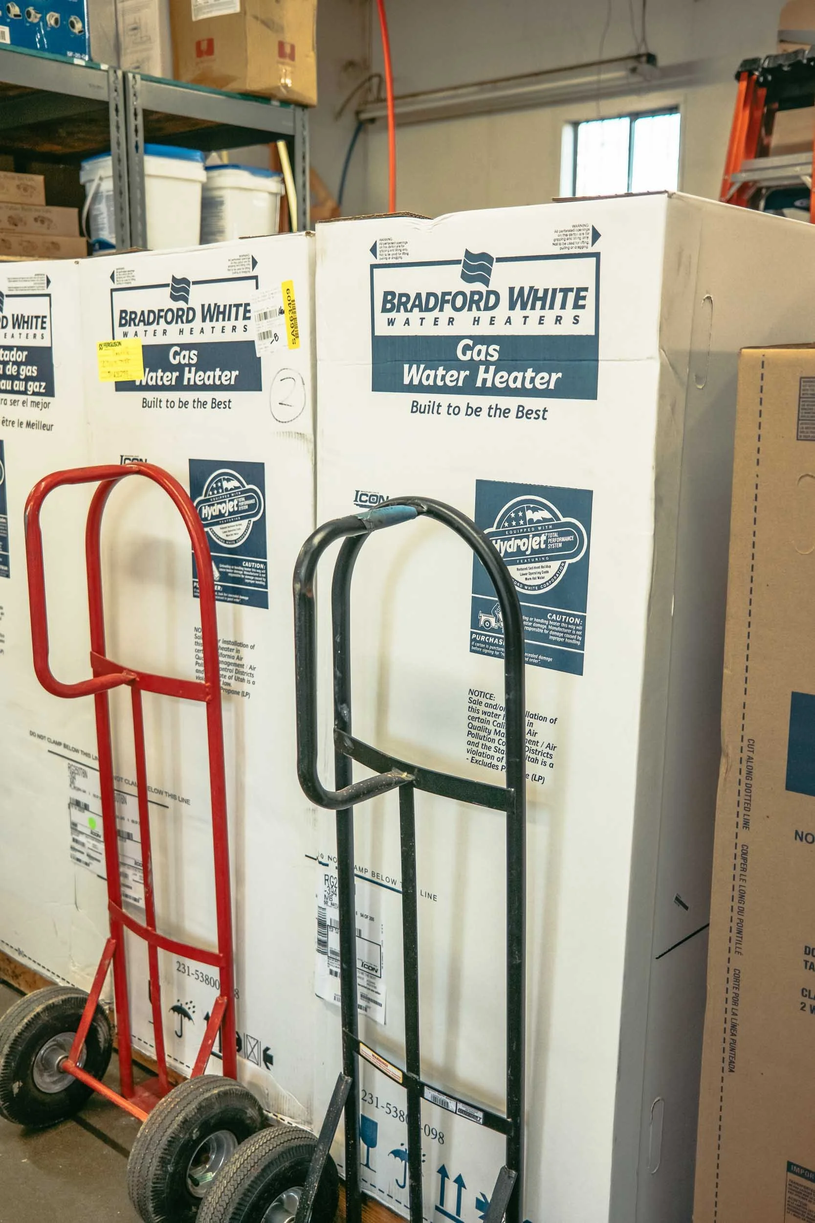 Two Bradford White water heaters in boxes, with two hand trucks in front of them, one red and one black, in a warehouse.