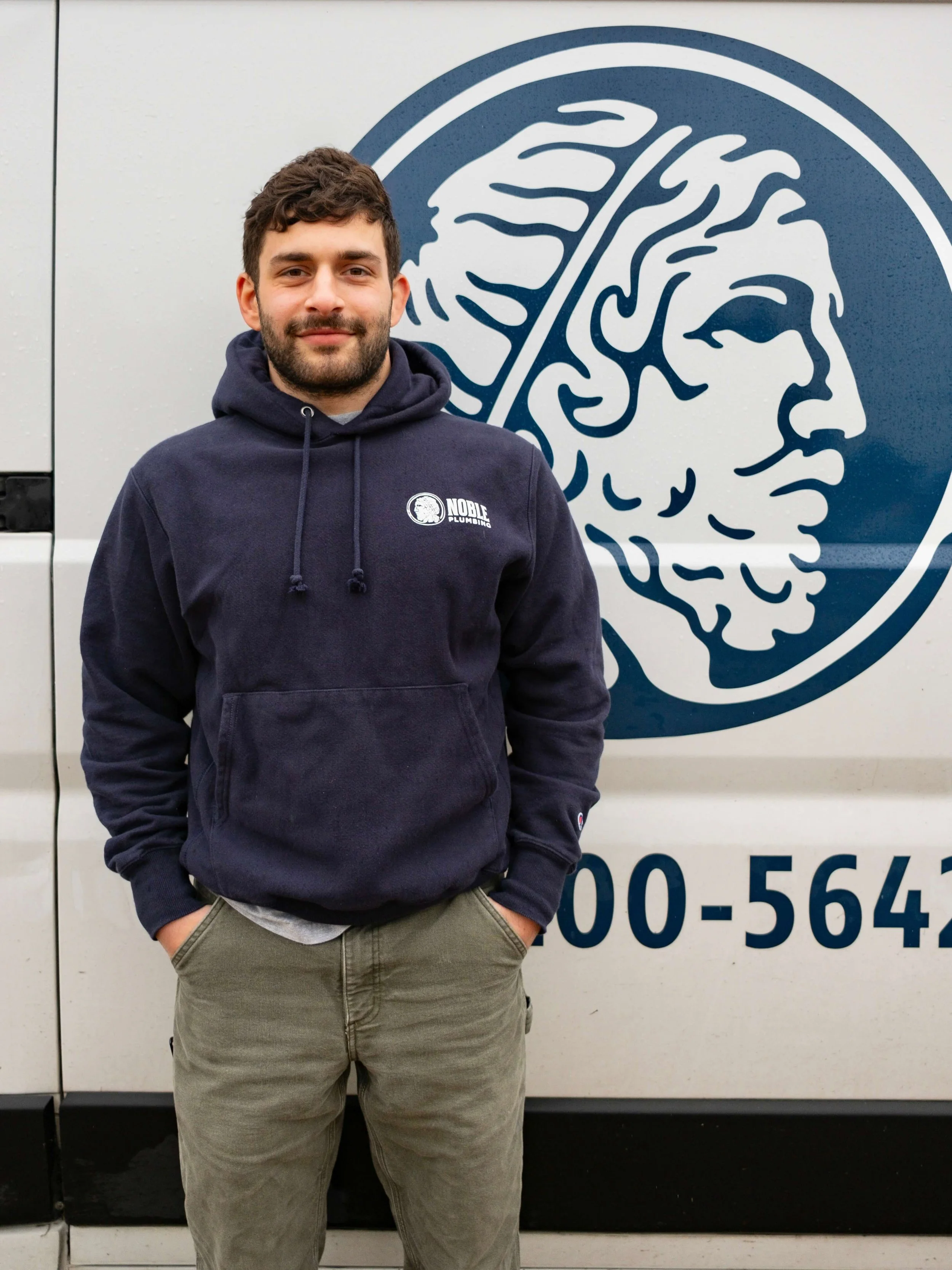 A smiling man with dark hair and beard, wearing a navy blue t-shirt that says Noble Plumbing, standing outdoors with tall grass and trees in the background. Joe is a Noble Plumbing owner.