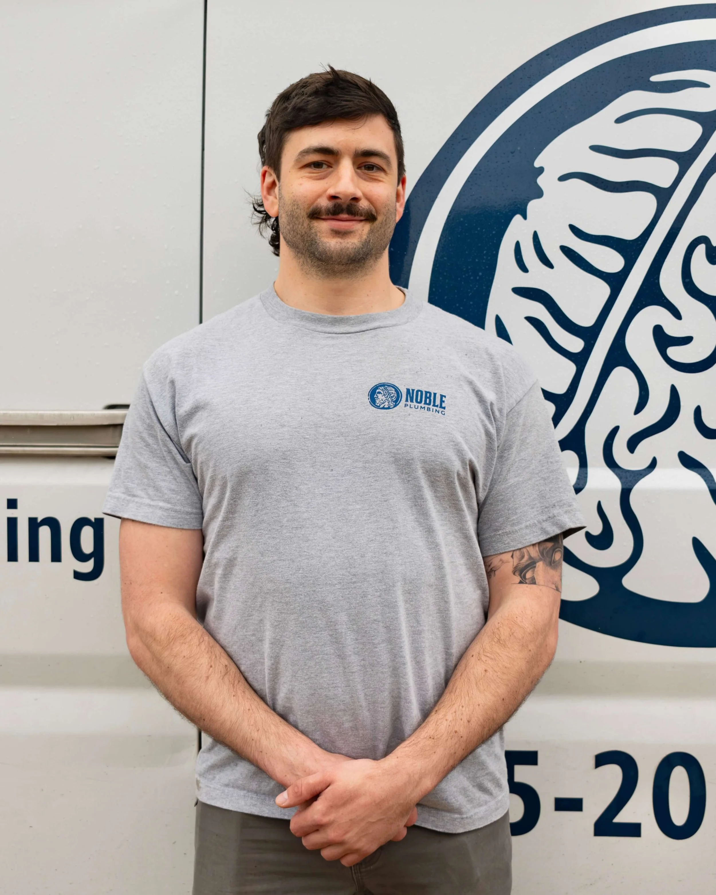 A man with dark hair, a mustache, and a beard wearing a blue sleeveless shirt with 'Noble Plumbing' written on it, standing outdoors in a green field with mountains and a blue sky in the background.