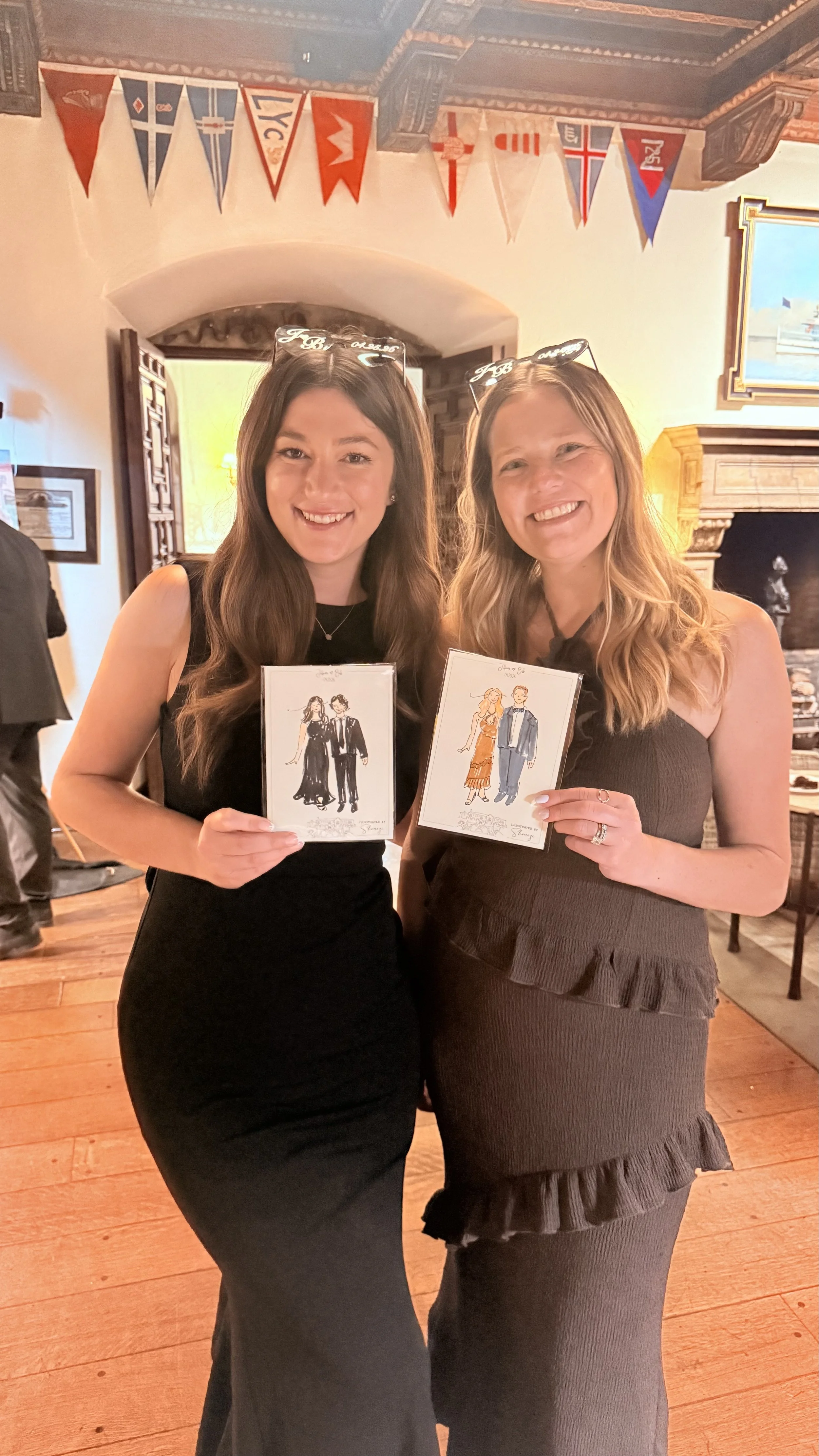 Two women in black and brown dresses standing inside a room with wooden floors, holding sketches of formal wear, smiling at the camera, with colorful nautical flags hanging from the ceiling behind them.