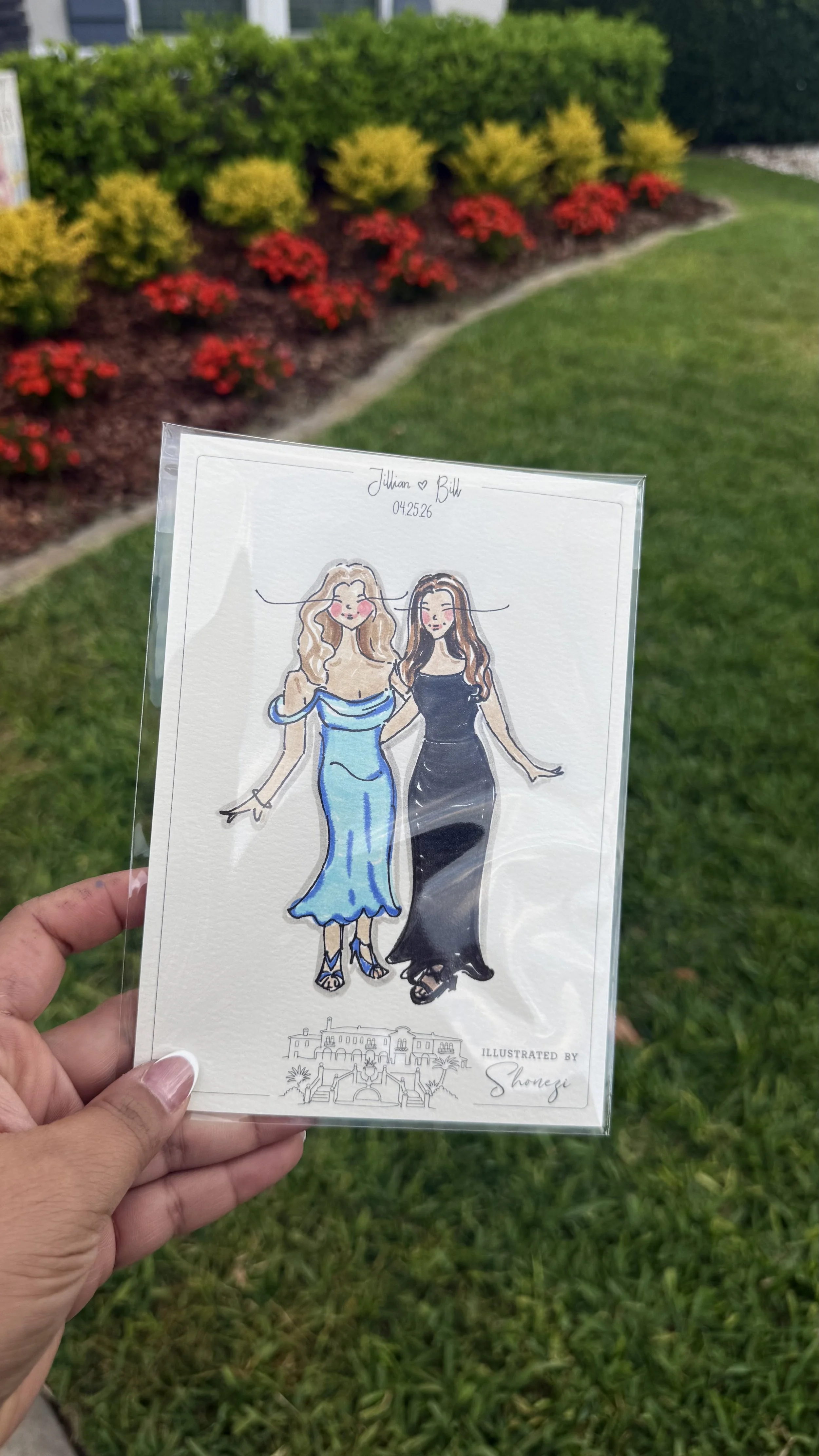 A hand holds a drawing of two women in elegant dresses, with a green lawn and colorful shrubbery in the background.