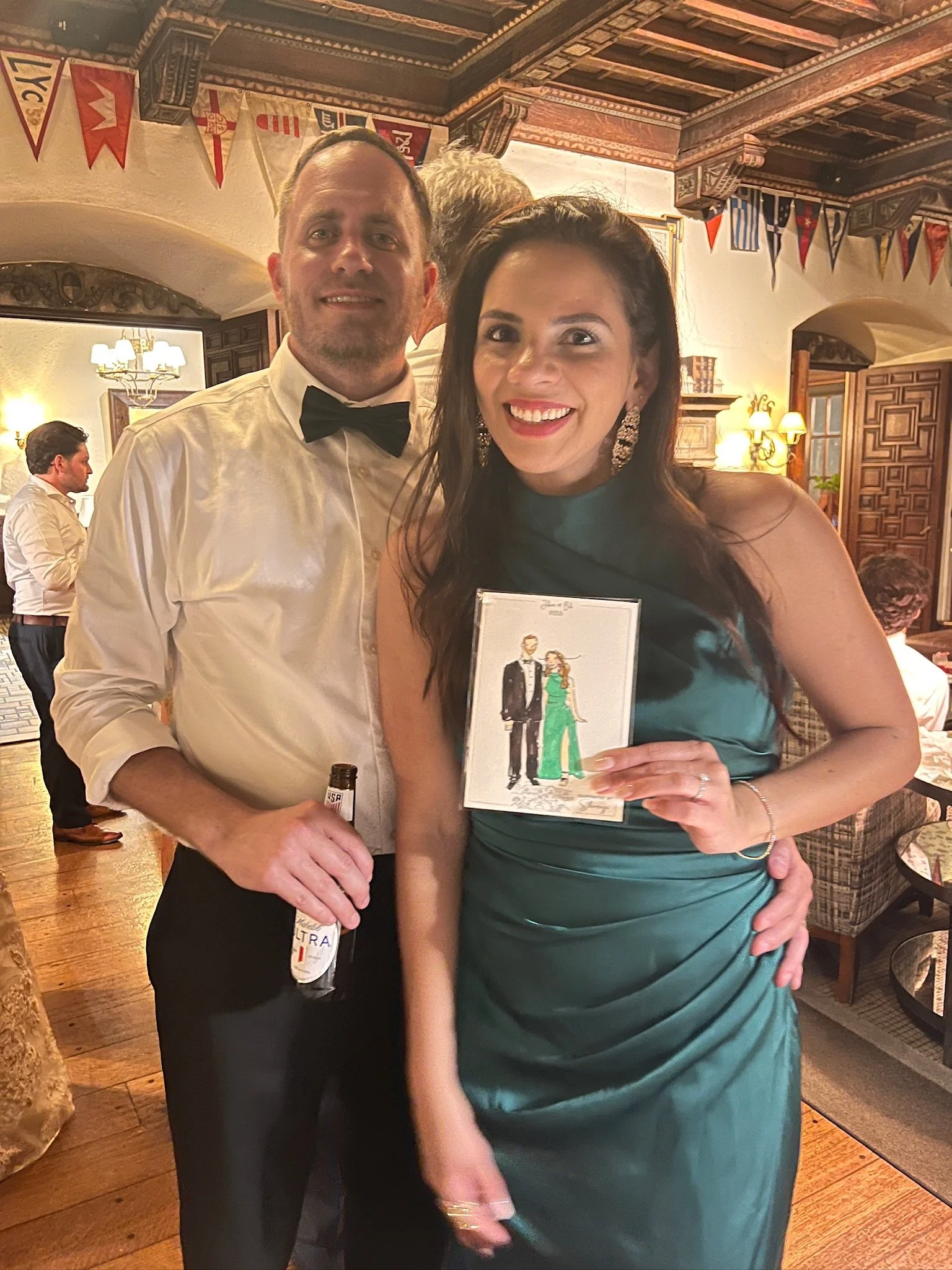 A smiling couple at a festive event, dressed in formal attire, with the man wearing a white shirt and black bow tie, and the woman wearing a teal dress and large earrings, holding a card featuring a cartoon illustration of a man in a tuxedo and a wom