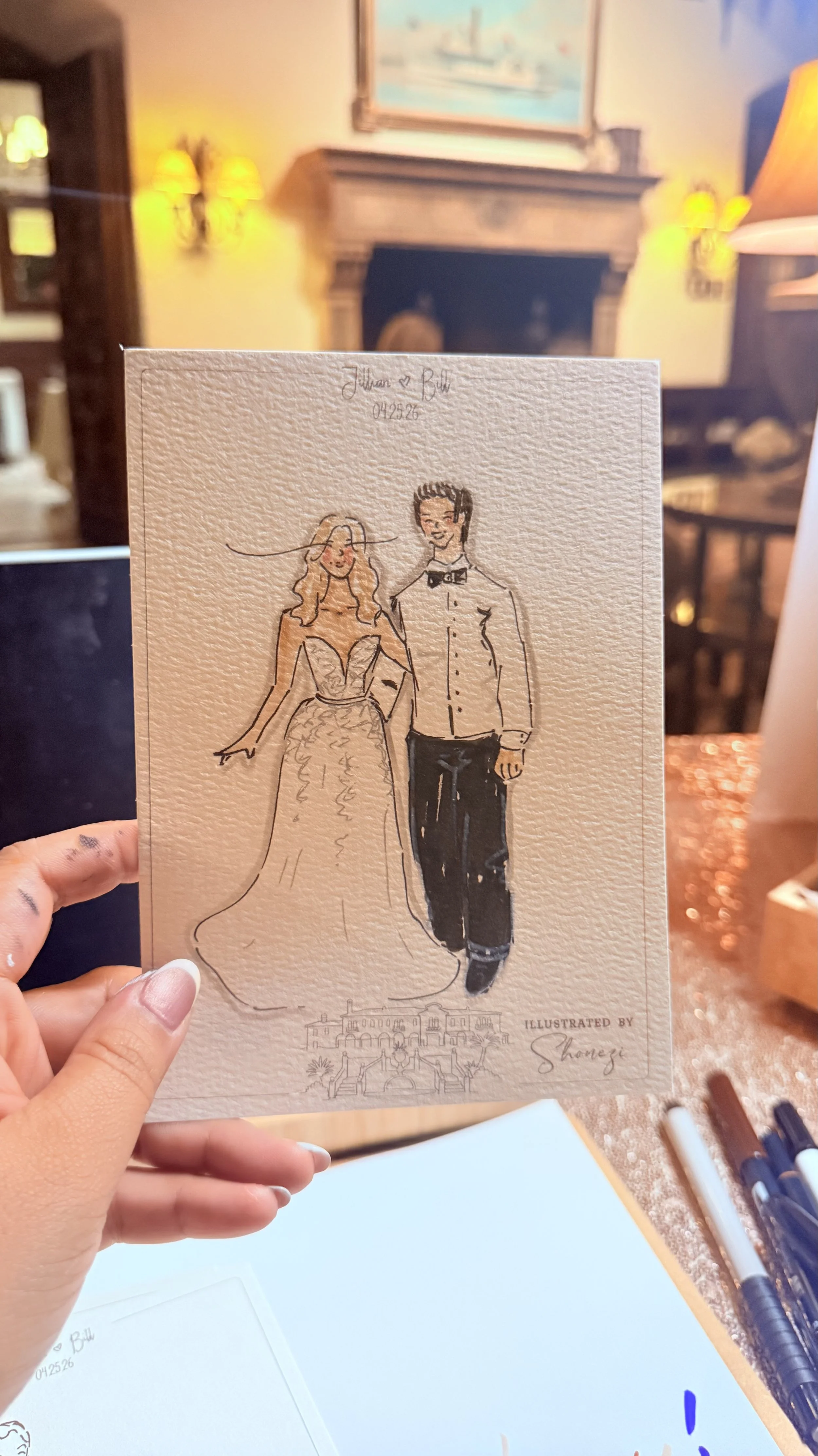 Hand-drawn illustration of a bride and groom in wedding attire, with the bride wearing a gown and the groom dressed in a tuxedo, held in front of a cozy living room with a fireplace.