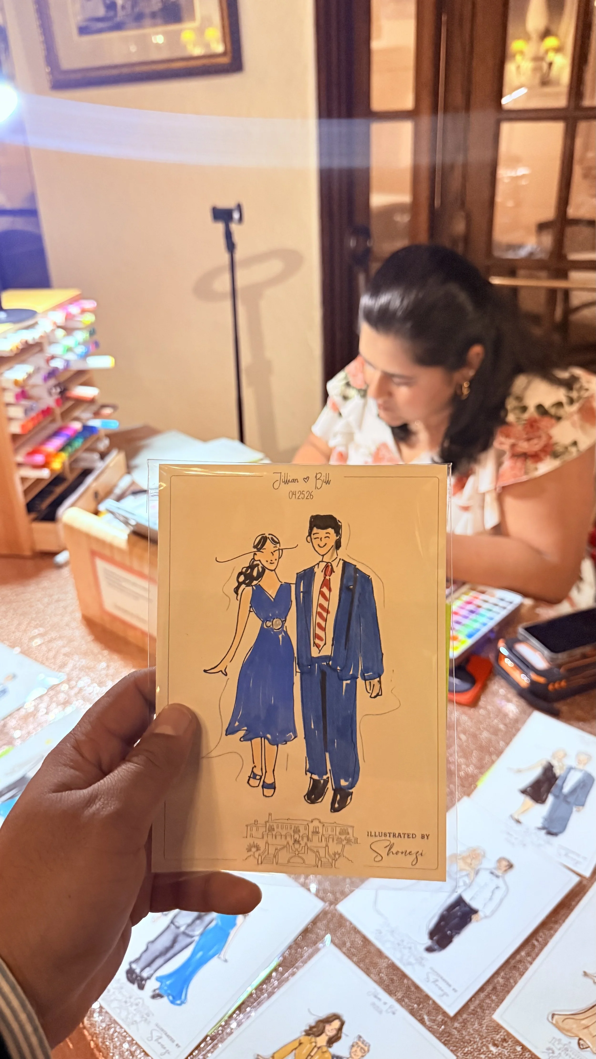 A hand holding a colorful illustration of a couple in formal attire, with a woman in a dress and a man in a suit and tie, in front of a woman sitting at a table with art supplies in the background.