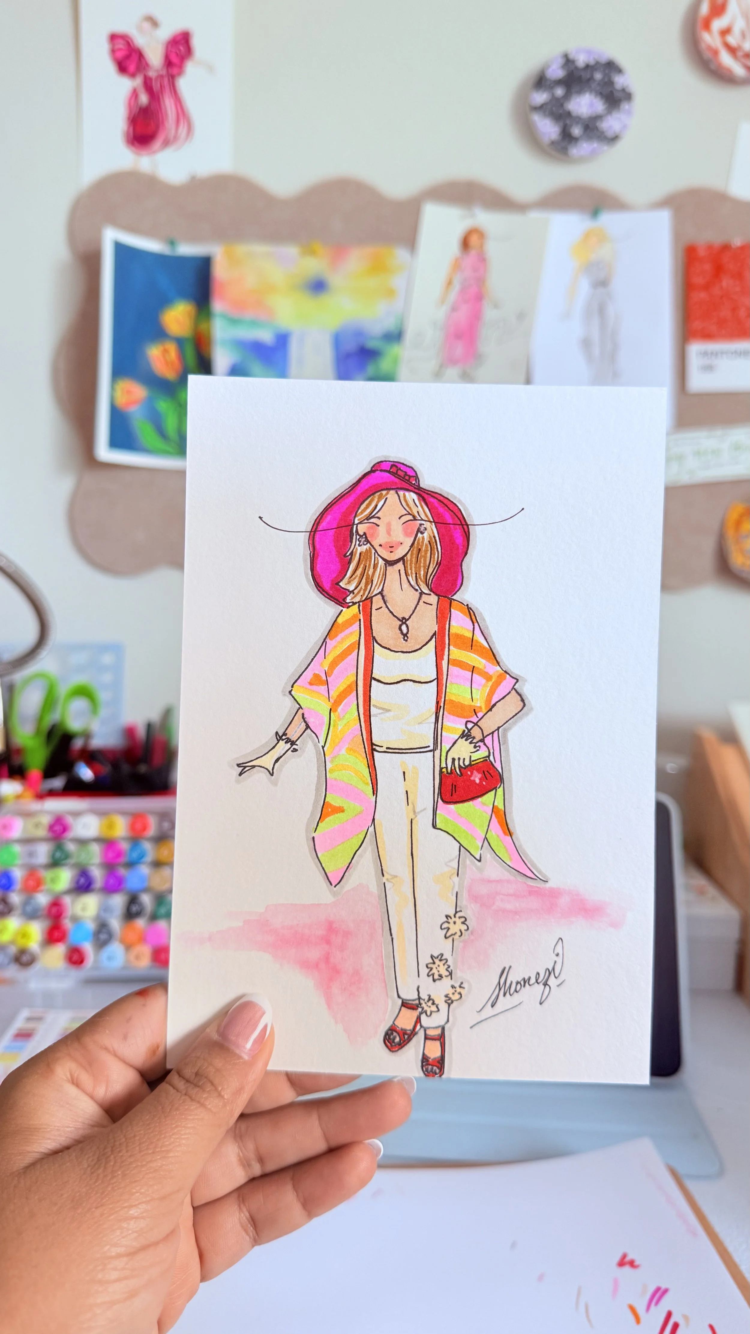 A hand holding a colorful fashion illustration of a woman in a pink hat, striped shawl, beige pants, high heels, and jewelry. The background shows an art display with drawings and paintings on a bulletin board.