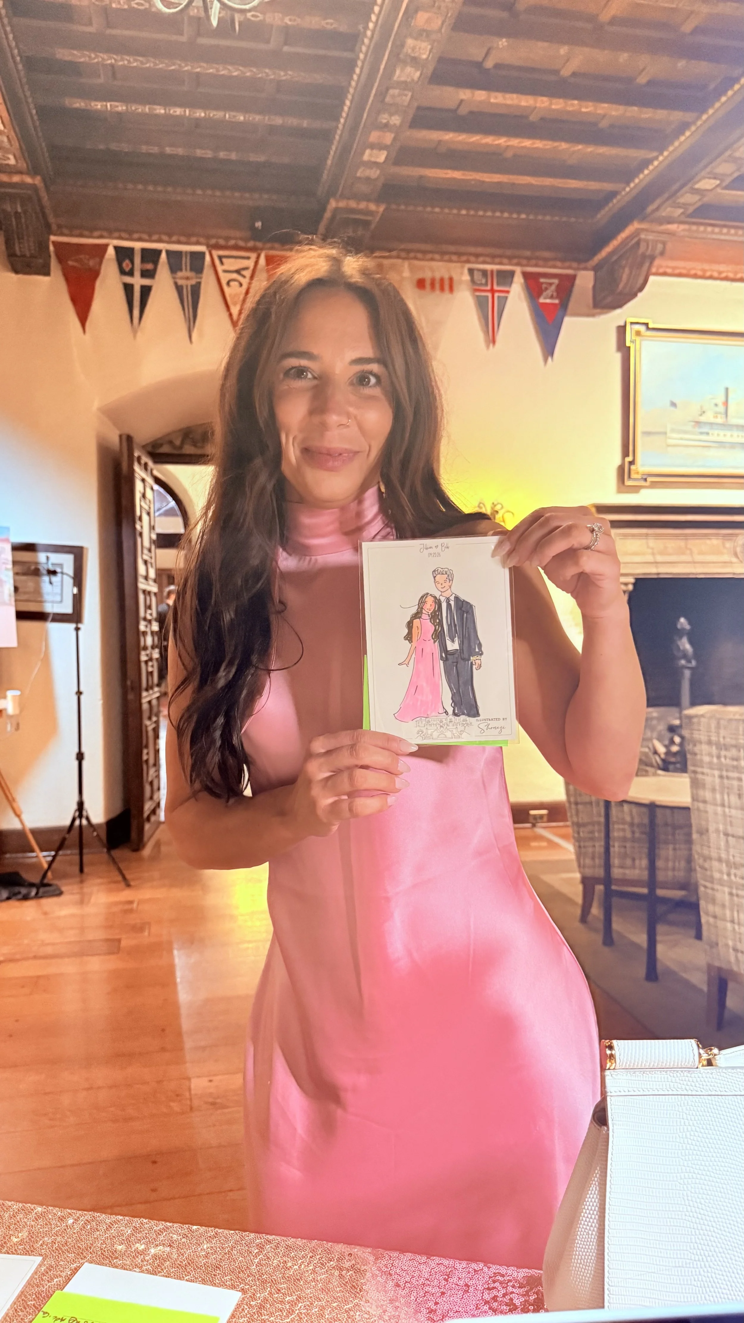 Woman in pink satin dress holding a drawing of a man and woman dressed in formal attire, standing in a warmly lit room with wooden ceiling, flags, and a fireplace.