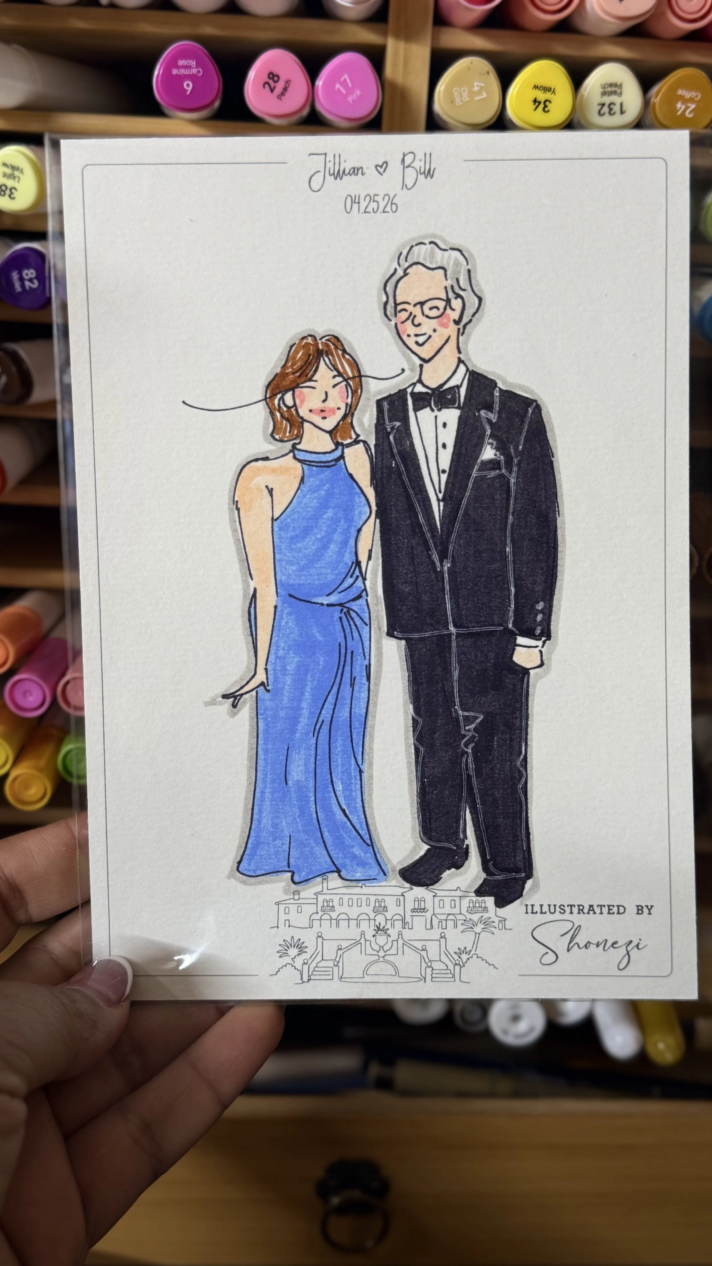 A hand holding a drawing of a couple dressed in formal attire, with the woman in a blue gown and the man in a tuxedo. The background shows shelves with colorful markers.