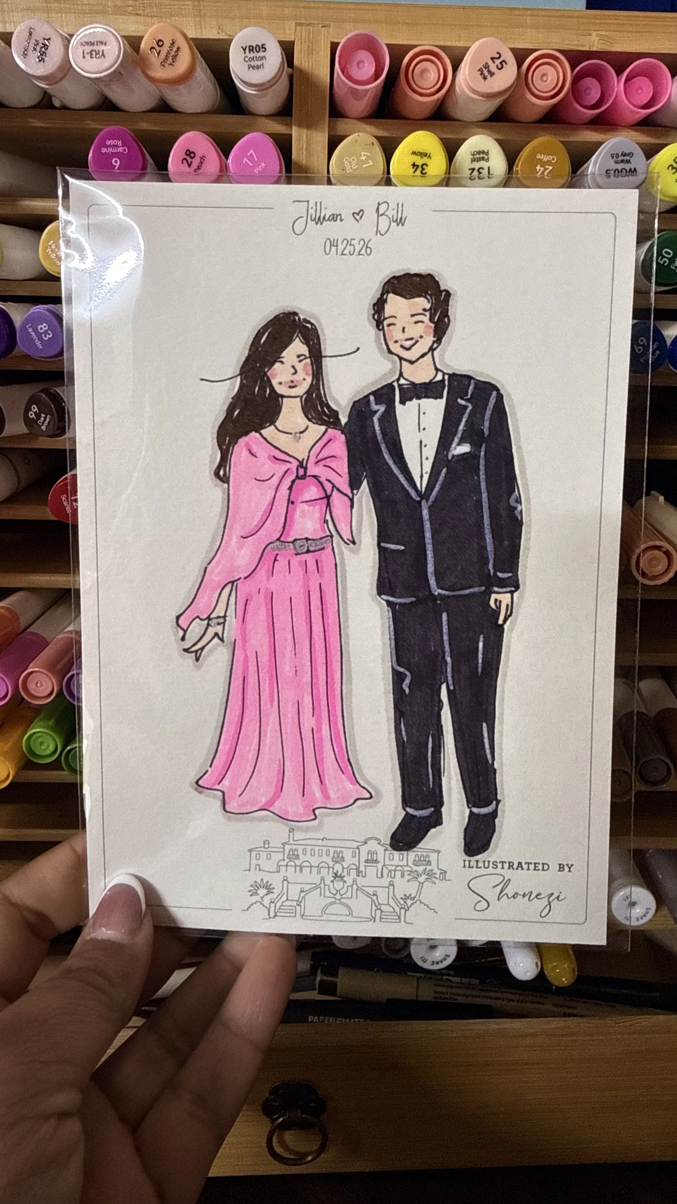 Hand-drawn illustration of a couple dressed in formal attire, with a woman in a pink gown and a man in a black tuxedo, standing together, on a white sheet of paper held in front of a box of colorful markers.