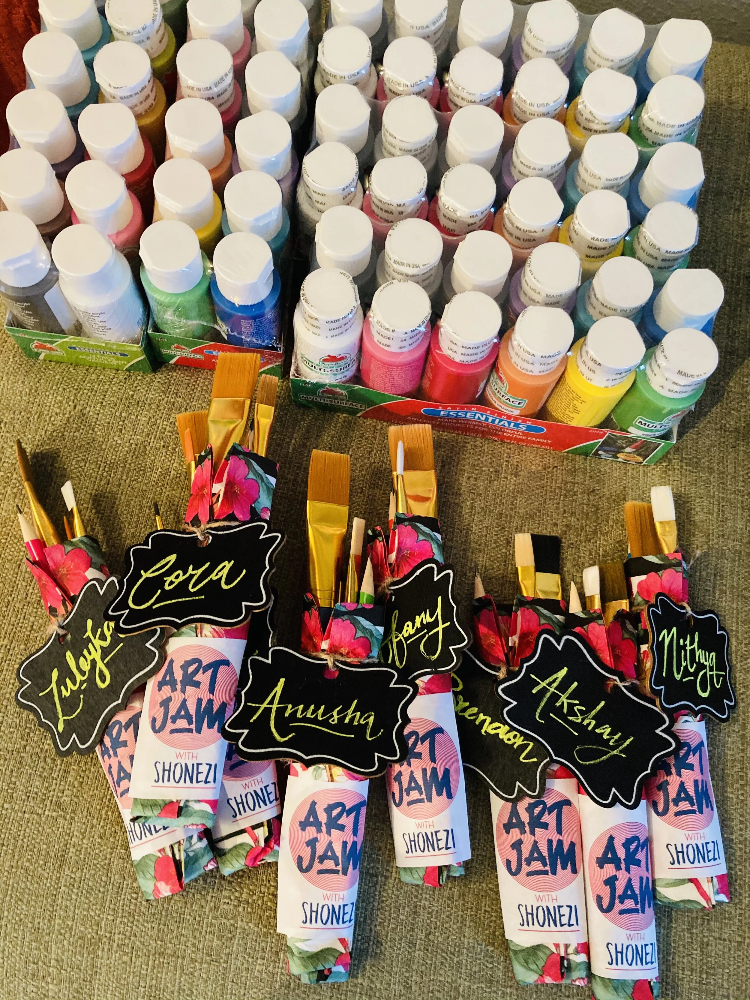 Colorful bottles of paint arranged in a box, with labeled art supplies wrapped in floral paper, each tag bearing a name such as Alek, Cora, Anusha, Samy, Akshat, and nothing.