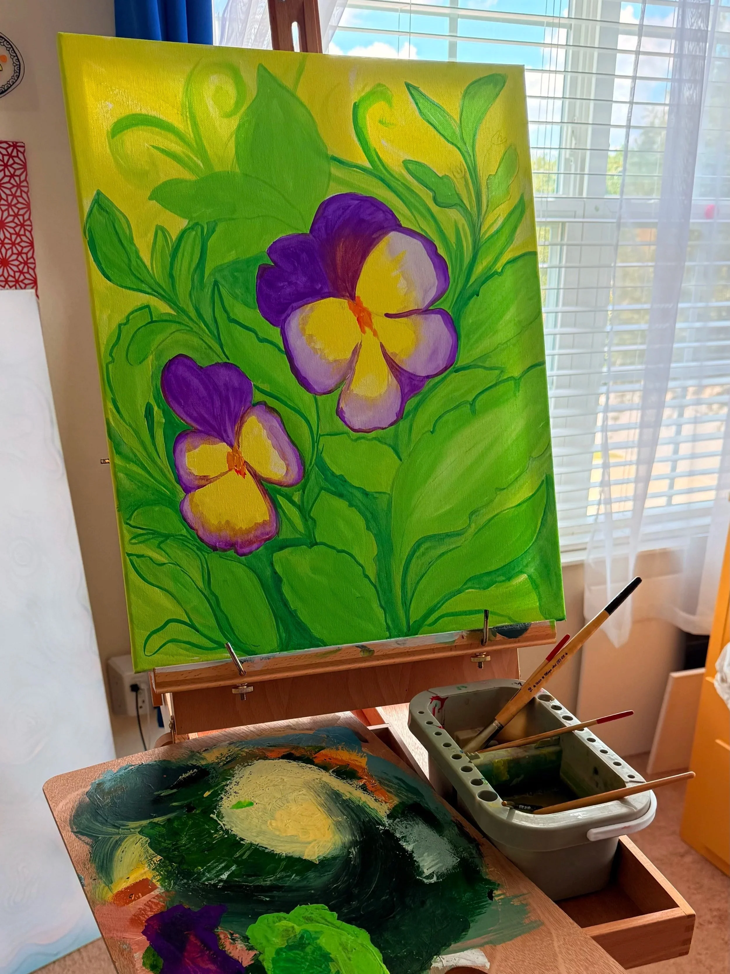 A colorful painting of purple and yellow flowers with green leaves on a yellow background, set on an easel in a sunlit room.