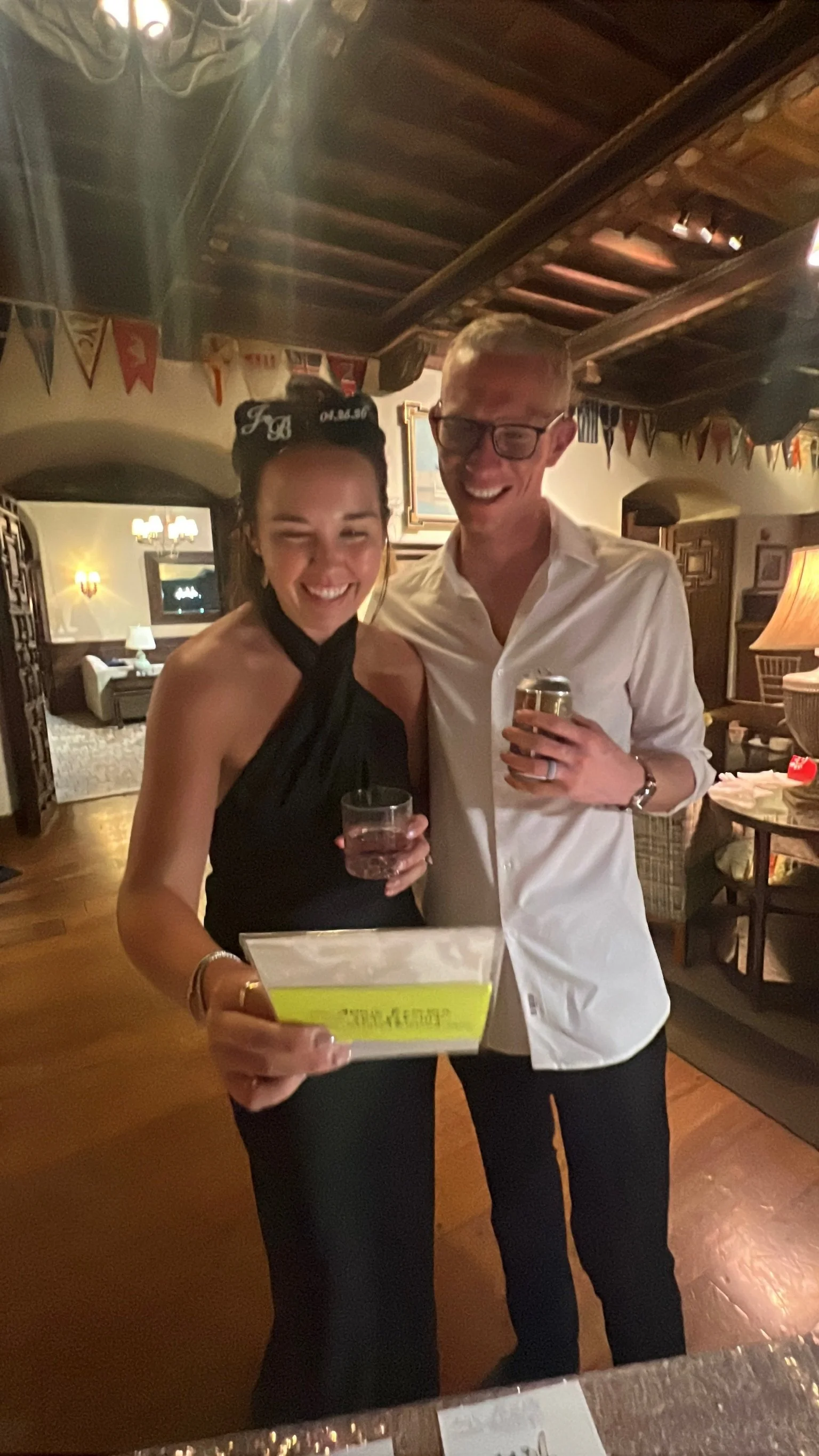 Two people, a woman and a man, smiling and posing for a photo indoors. The woman is wearing a black dress and holding a drink and a yellow paper; the man is wearing a white shirt, glasses, and holding a drink. The background shows a cozy, warmly lit 