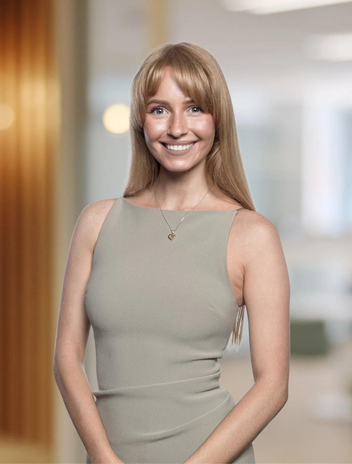 A smiling woman with strawberry blonde hair wearing a sleeveless gray dress and a necklace, standing indoors with a blurred background.