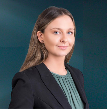 University or professional woman with long brown hair, wearing a black blazer and green blouse, posing against a dark teal background.