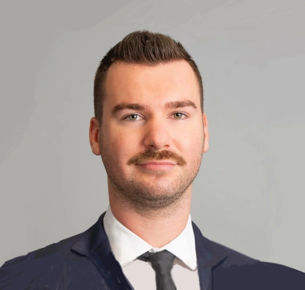 A young man with short brown hair, a mustache, wearing a black suit, white shirt, and black tie, standing against a light gray background.