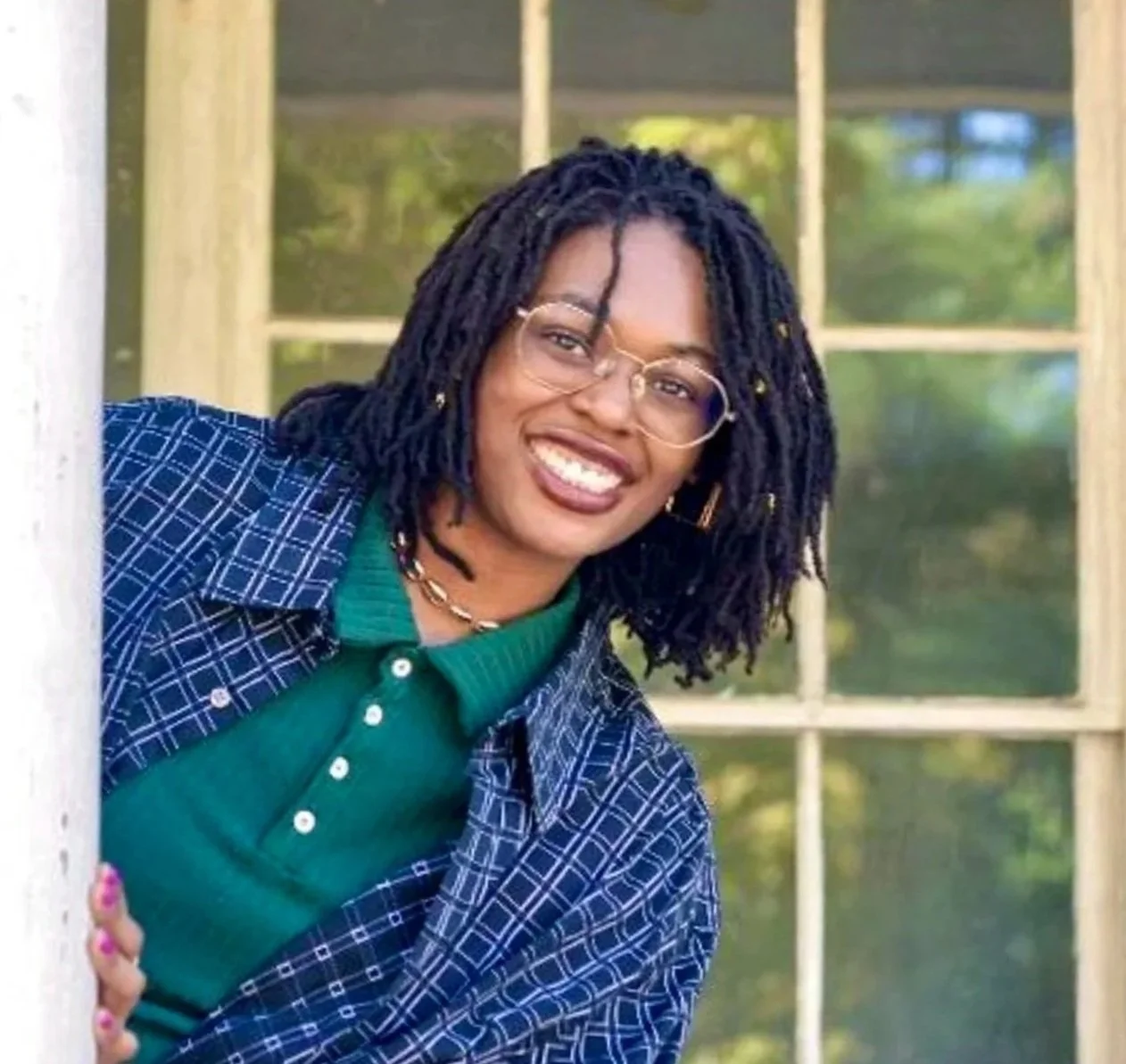 A woman with dark braided hair, glasses, and a big smile leaning out of a window, wearing a green shirt and a blue plaid jacket with a background of a window and greenery.