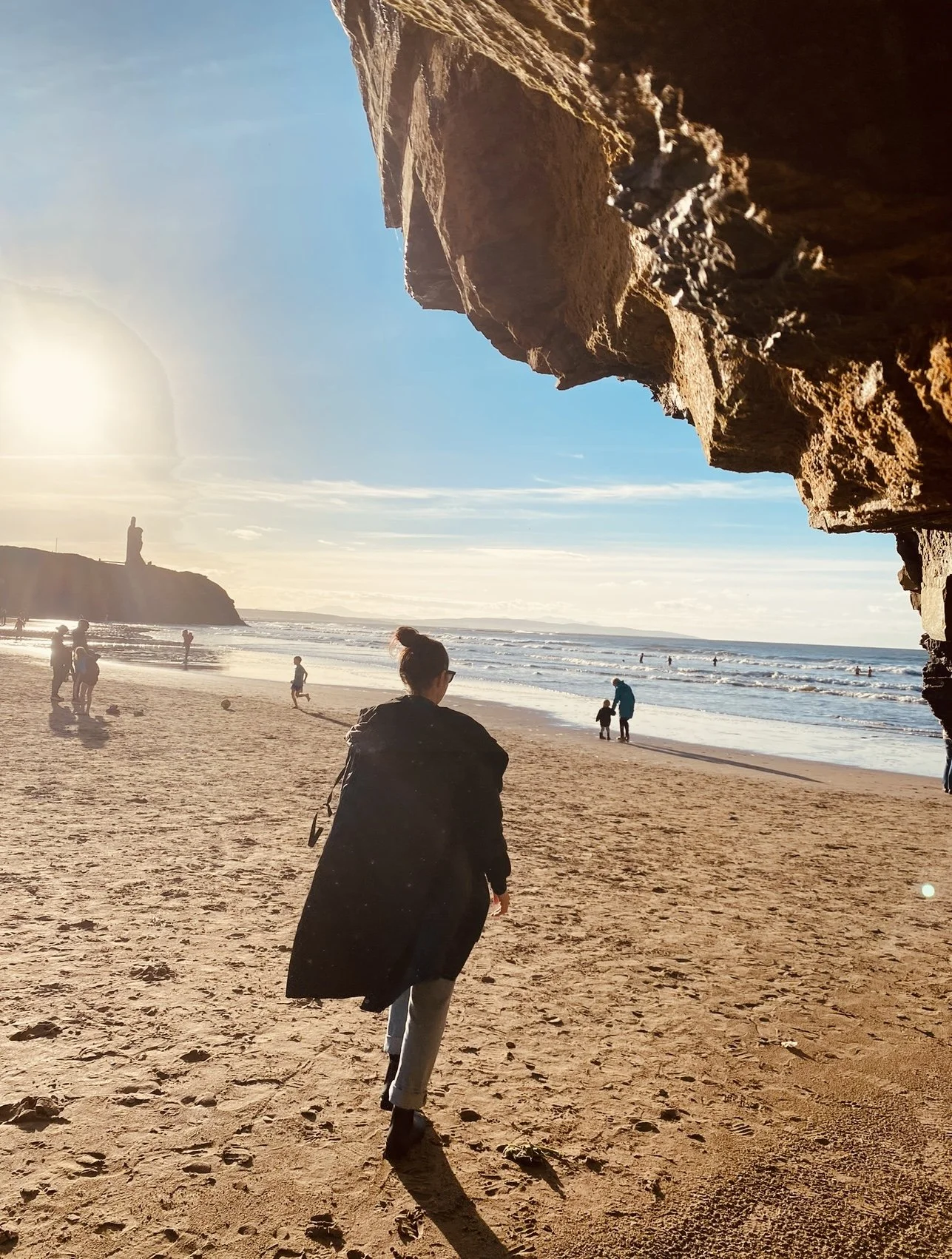 A woman walking on a sandy beach near the ocean, with a large rock formation in the foreground and a lighthouse on a cliff in the background.