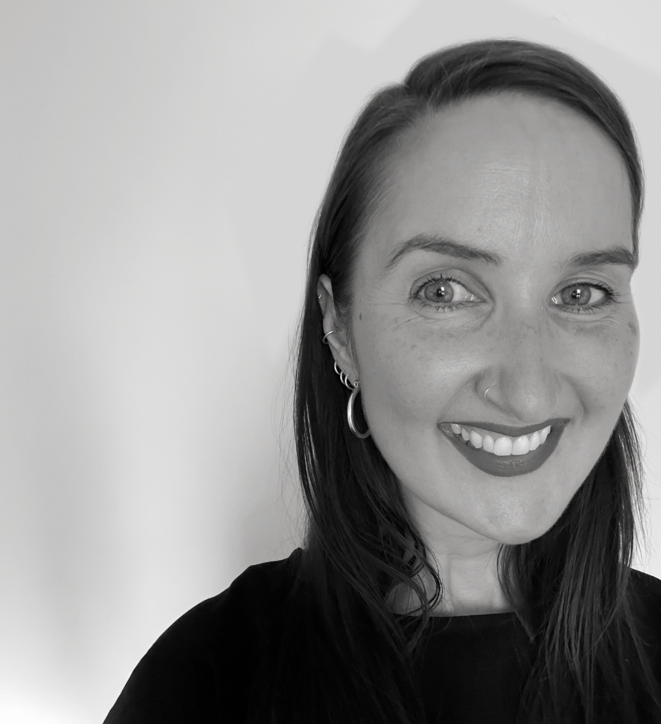 A black and white photo of a smiling woman with dark hair, wearing multiple earrings, a nose ring, and a dark top, standing against a plain light background.