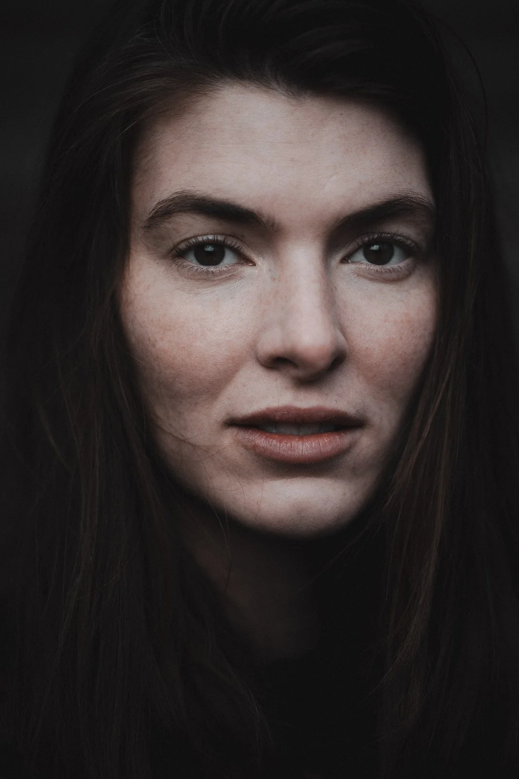 Nahaufnahme eines porzellanfarbenen Gesichts mit braunen Haaren, blauen Augen und natürlichen Lippen vor einem dunklen Hintergrund.