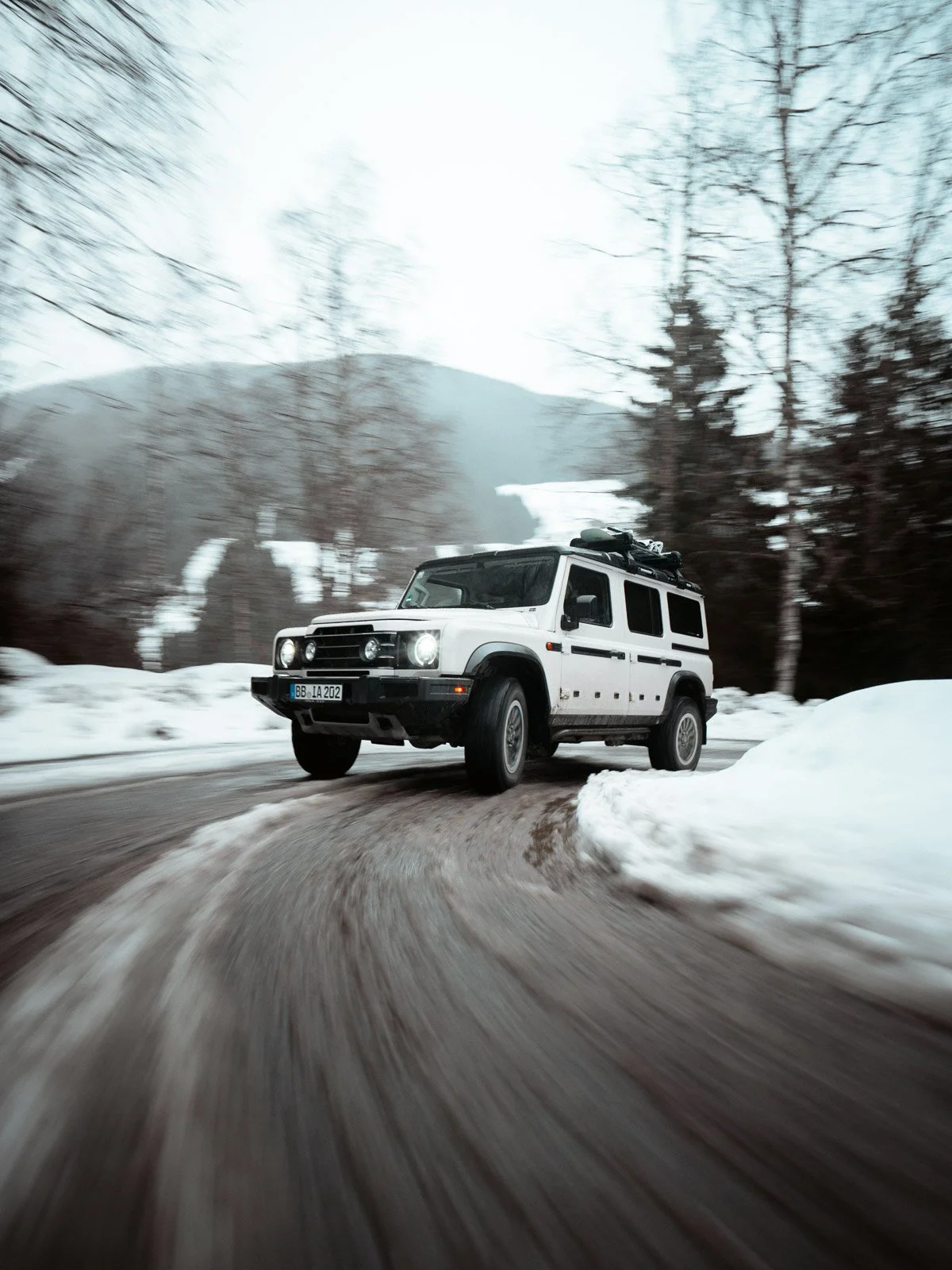 Ein weißer Geländewagen fährt auf einer kurvigen Straße im winterlichen Wald. Schnee liegt am Straßenrand, Bäume sind kahl, Berge im Hintergrund.