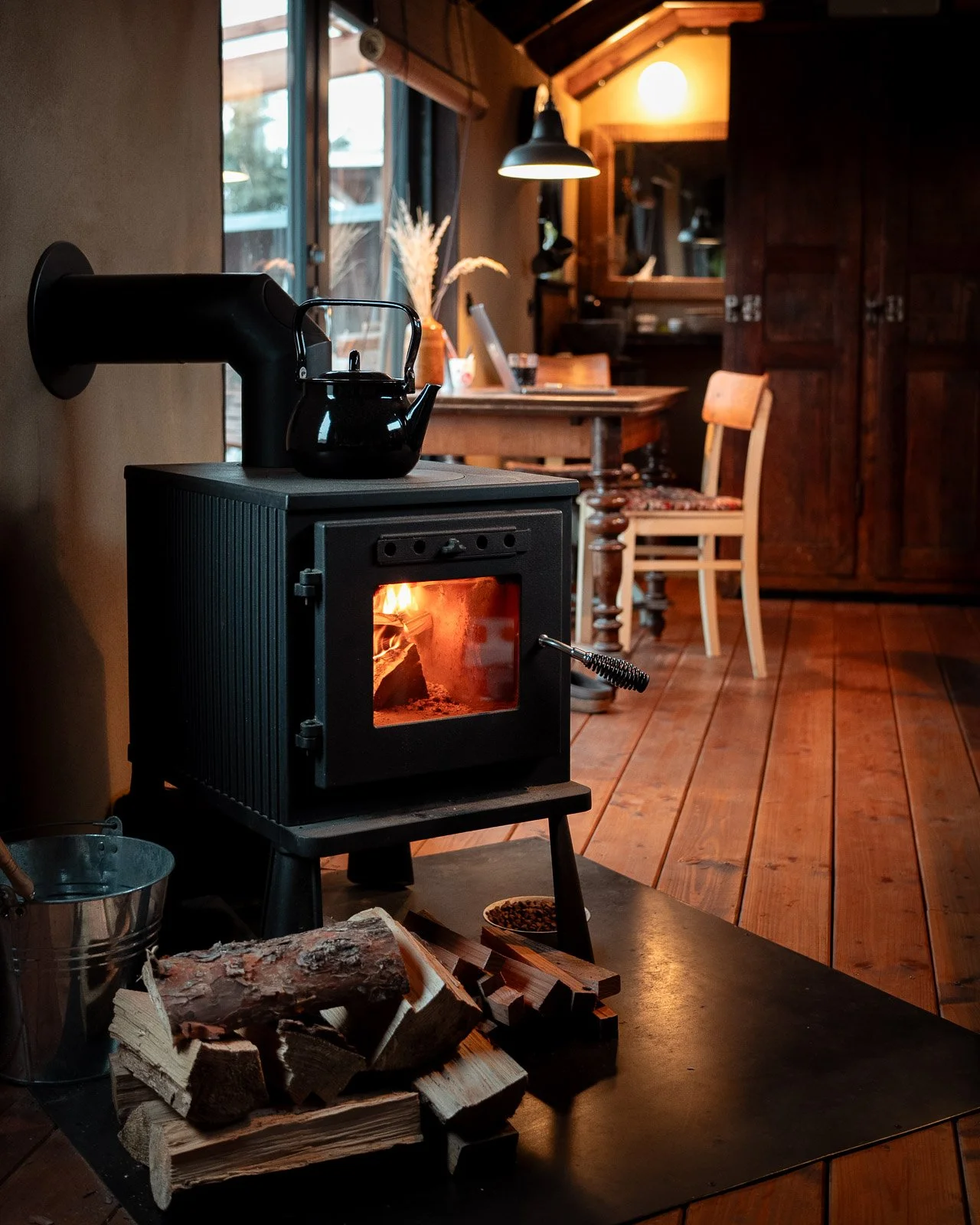 Ein Indoor-Ofen mit brennendem Feuer, auf dem eine schwarze Teekanne steht. Im Vordergrund liegen Holzscheite. Im Hintergrund ein Tisch, Stühle und eine Lampe in einem rustikalen Raum.