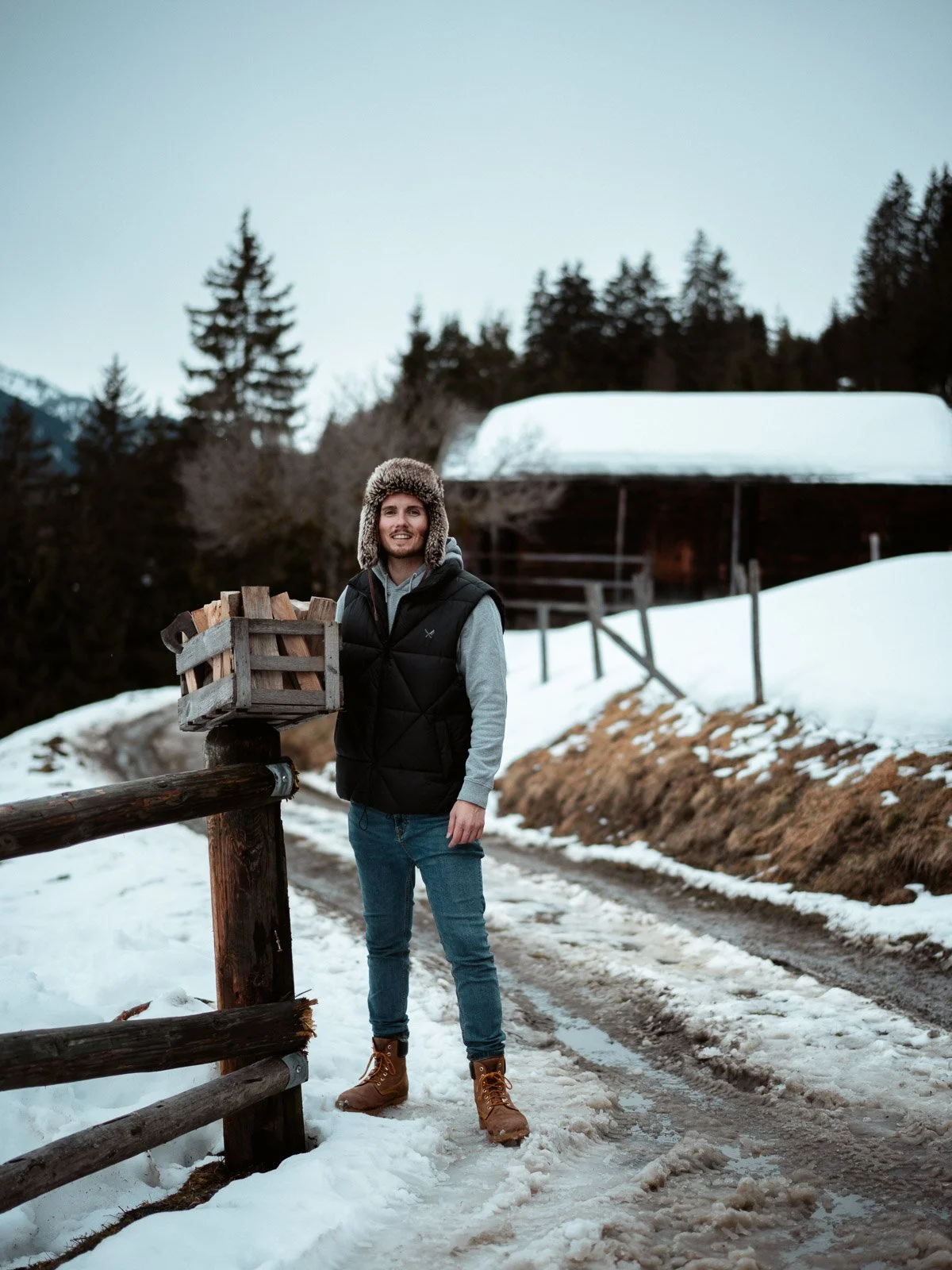 Ein Mann steht im Schnee neben einem Holzpoller, trägt eine Wintermütze und einen schwarzen Weste, im Hintergrund sind Bäume und ein Schneeschuppen sichtbar.