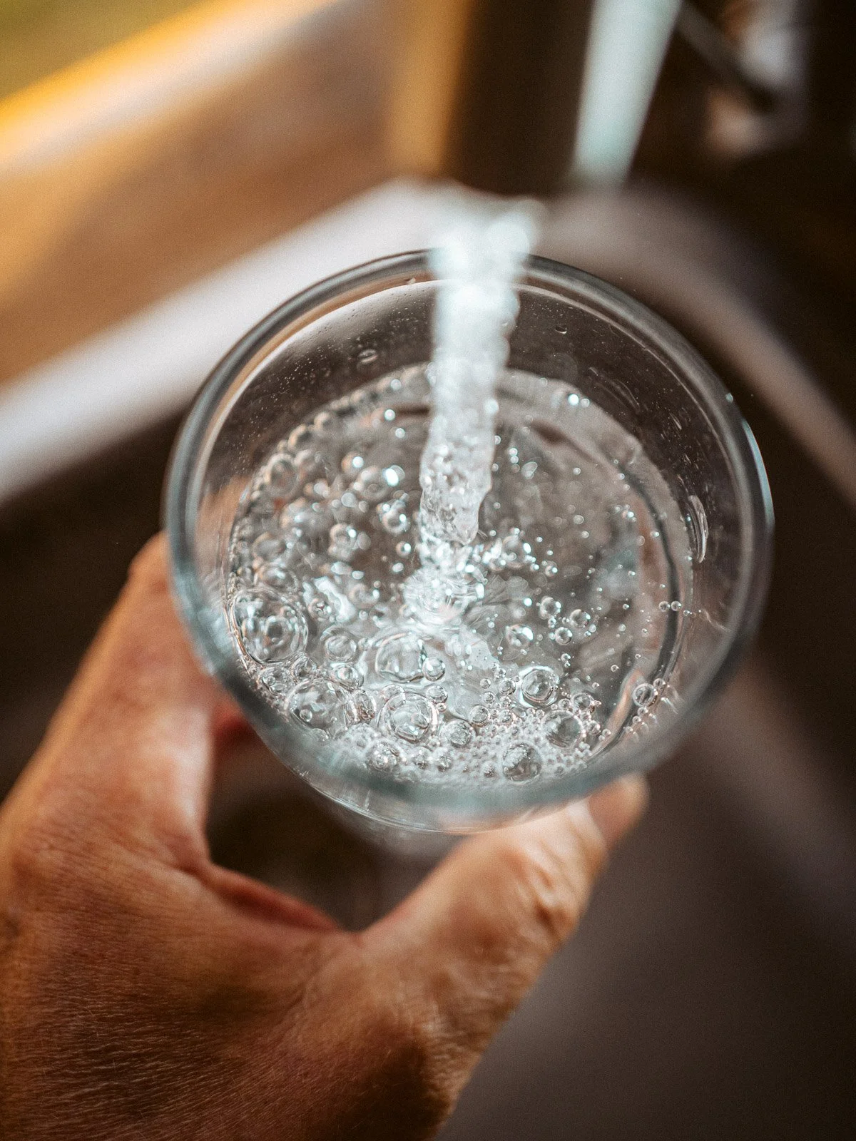 Nahaufnahme eines Glases, aus dem Wasser eingegossen wird, mit Bläschen an der Oberfläche, gehalten von einer Hand.