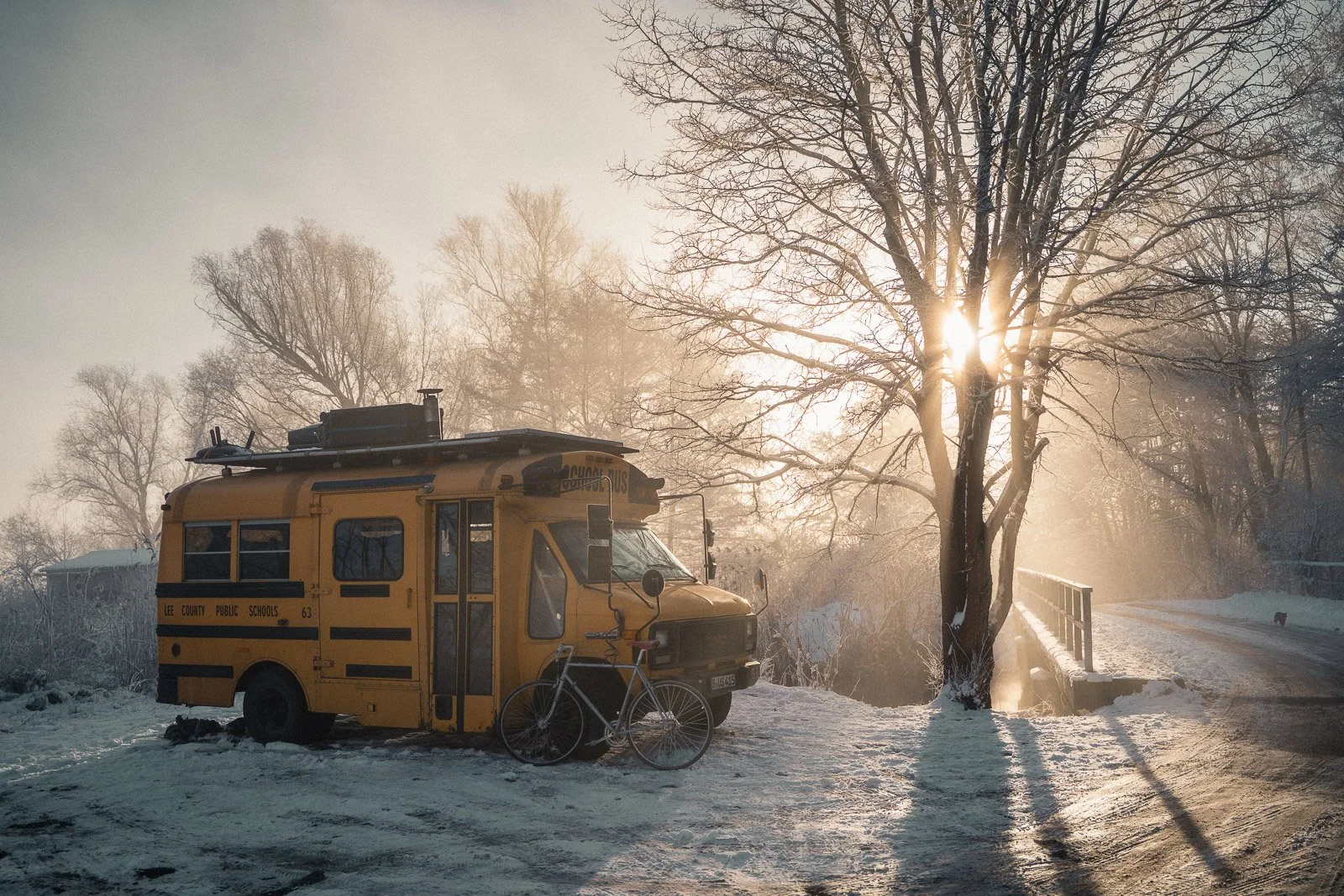 Ein gelber Schulbus steht im Schnee bei Sonnenaufgang, mit einem Fahrrad davor, in einer winterlichen Landschaft mit mehreren Bäumen und einem kleinen Fluss oder Bach