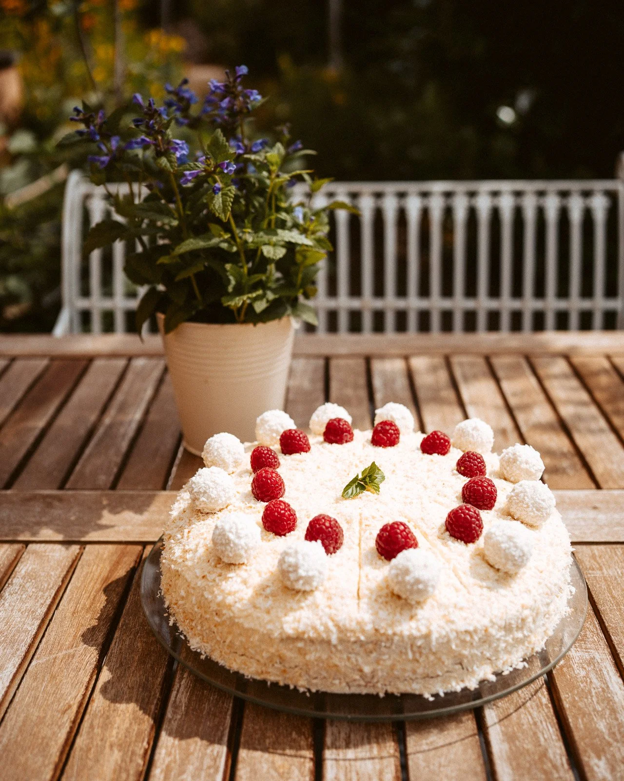 Ein Kuchen mit weißen Kokosraspeln, roten Himbeeren und einem kleinen grünen Blatt darin, auf einem Holztablett, im Freien bei Nacht.