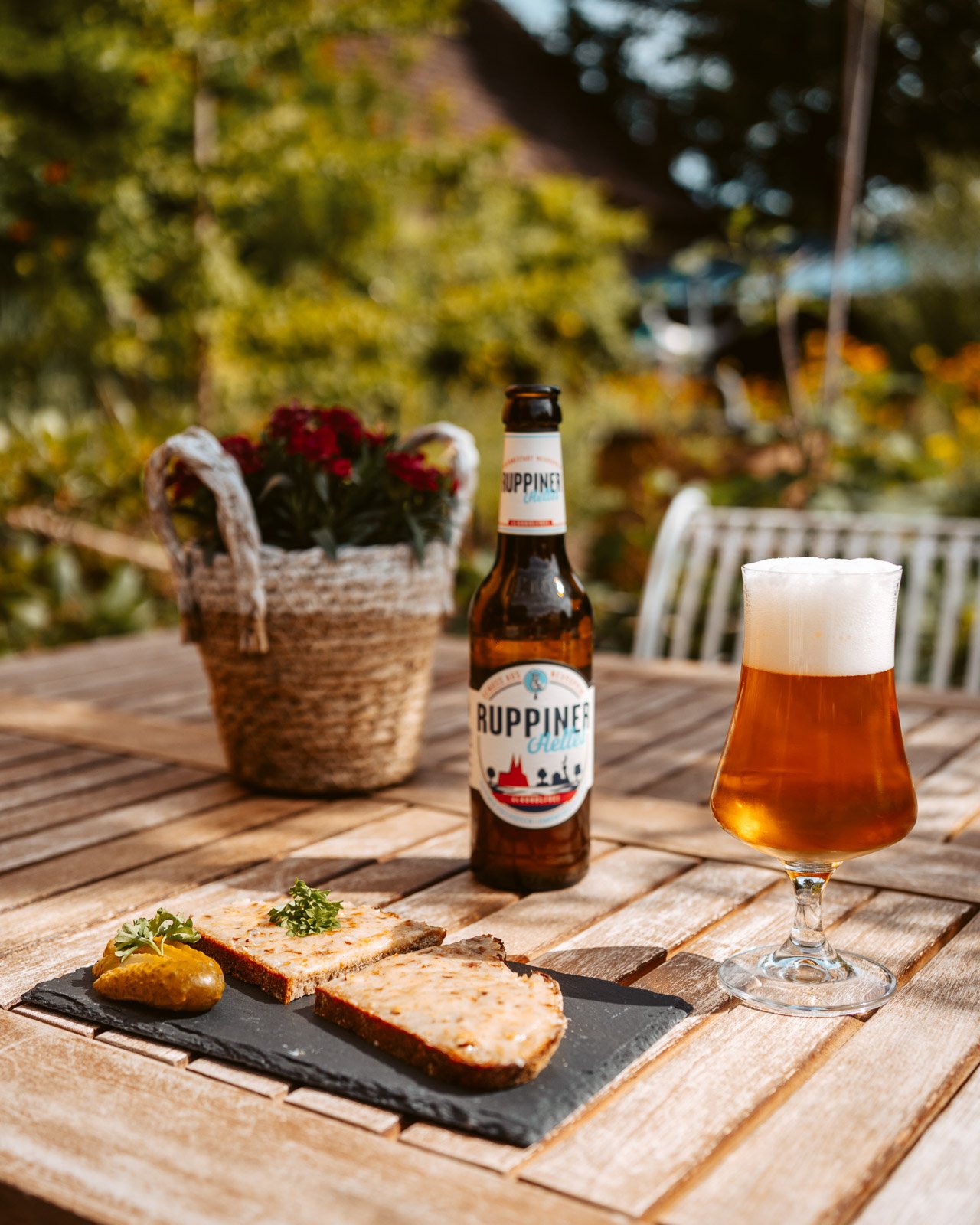 Ein Tisch im Freien mit einer Flasche Ruppiner Bier, einem Bierglas und zwei belegten Broten mit Senf und Kräutern, im Hintergrund eine Blumenvase. Naturgarten mit Bäumen im Sonnenlicht.