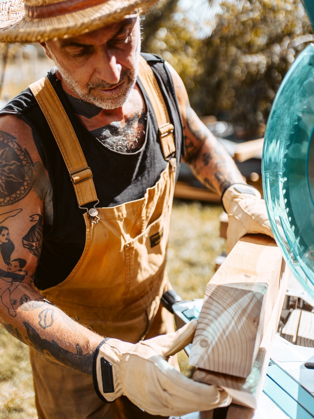 Ein gestikulierender männlicher Handwerker trägt braune Arbeitshandschuhe, eine Sonnenbrille, einen Strohhut, schwarze T-Shirt und braune Arbeitskleidung und arbeitet an einem Holzprojekt im Freien an einem sonnigen Tag.