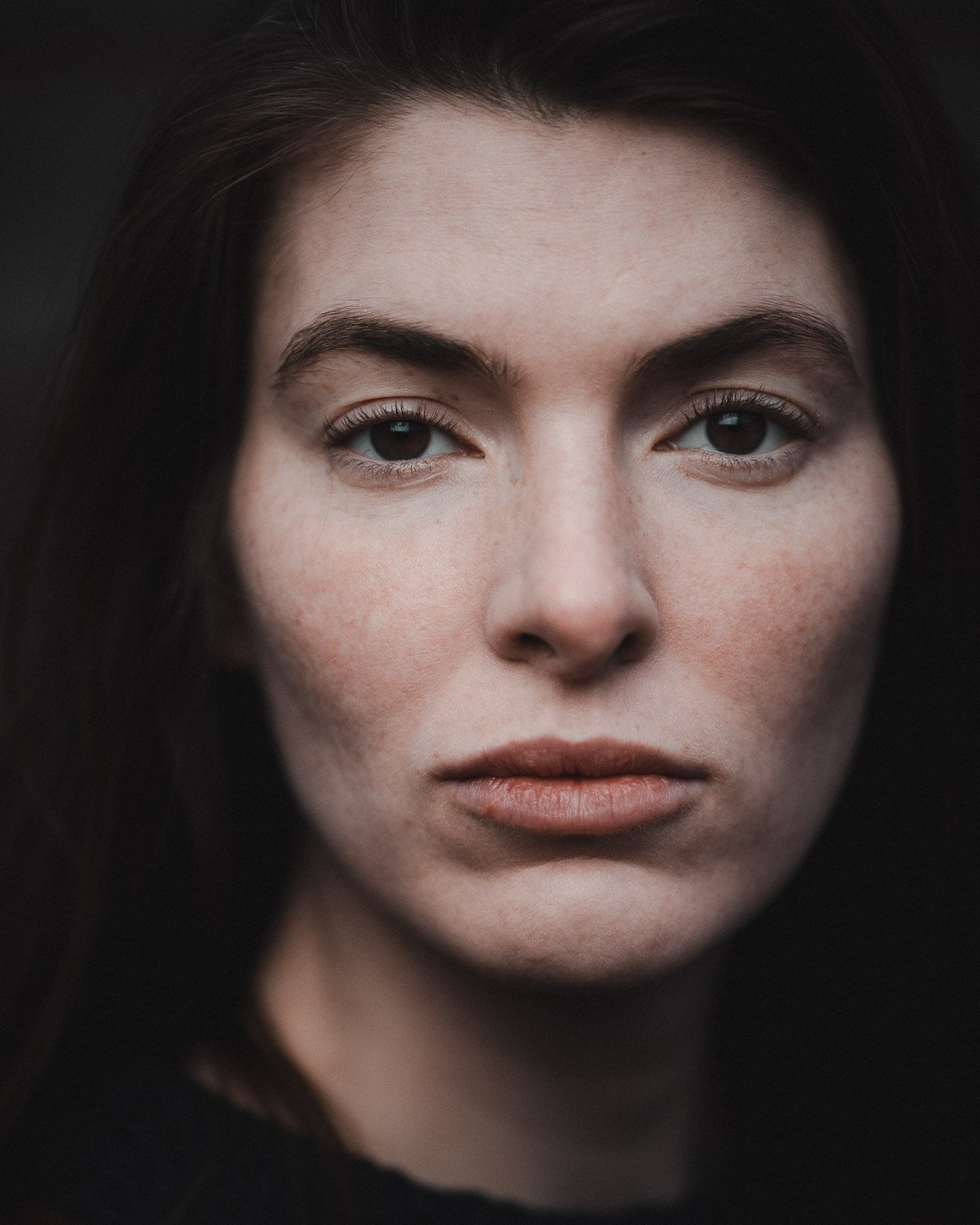 Nahaufnahme eines jungen Gesichts einer Frau mit dunklem Haar und blauen Augen, neutralem Ausdruck, helle Haut, dunklem Hintergrund.