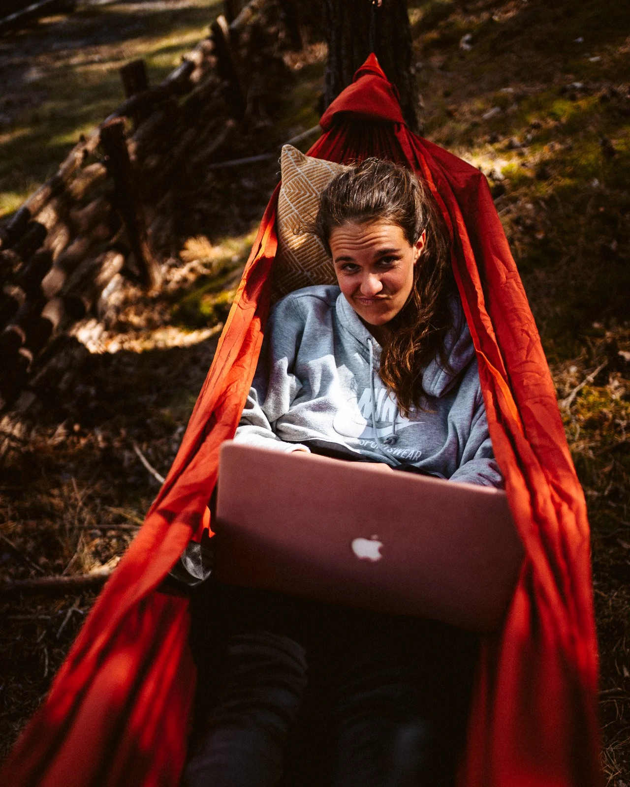 Junge Frau liegt in einer roten Hängematte im Wald, mit einem Laptop auf ihrem Schoß, trägt ein graues Hoodie, schaut skeptisch in die Kamera.