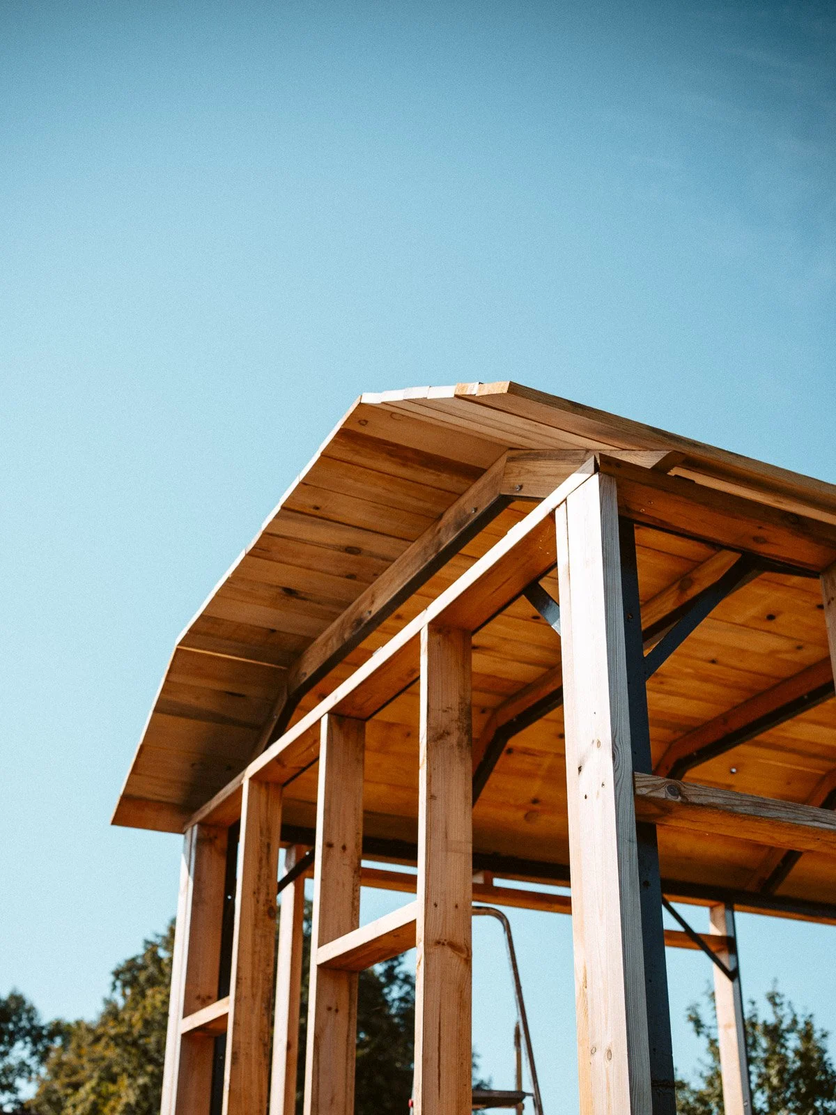 Holzrahmen für ein kleines Baumhaus oder Spielhaus, das im Bau ist, mit einer Holzbodenplatte und einem Dach aus Holzplanken, gegen den blauen Himmel. Im Hintergrund sind Bäume sichtbar.