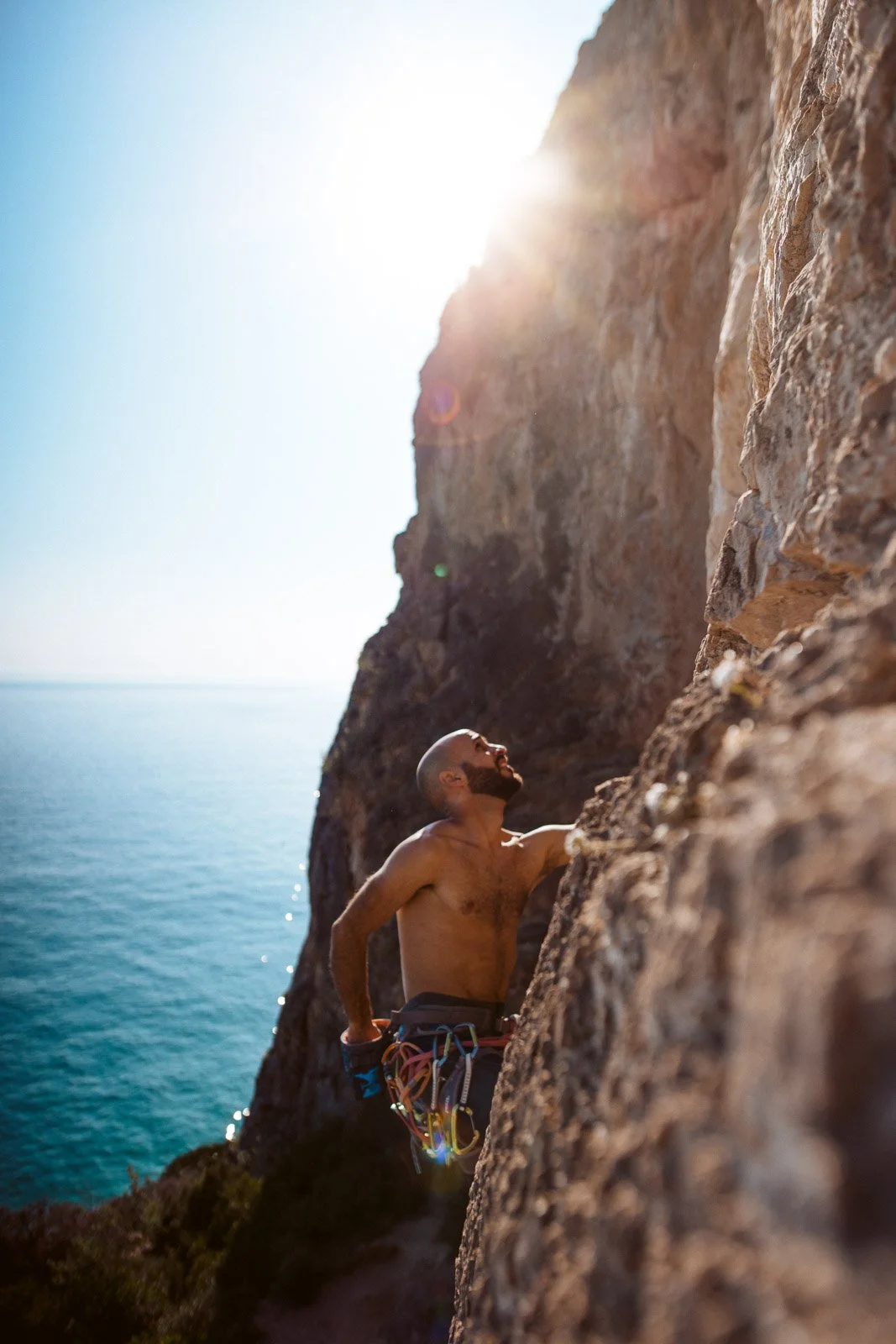 Männlicher Kletterer ohne Hemd klettert an einer steilen Felswand mit Blick auf das Meer bei Sonnenschein.