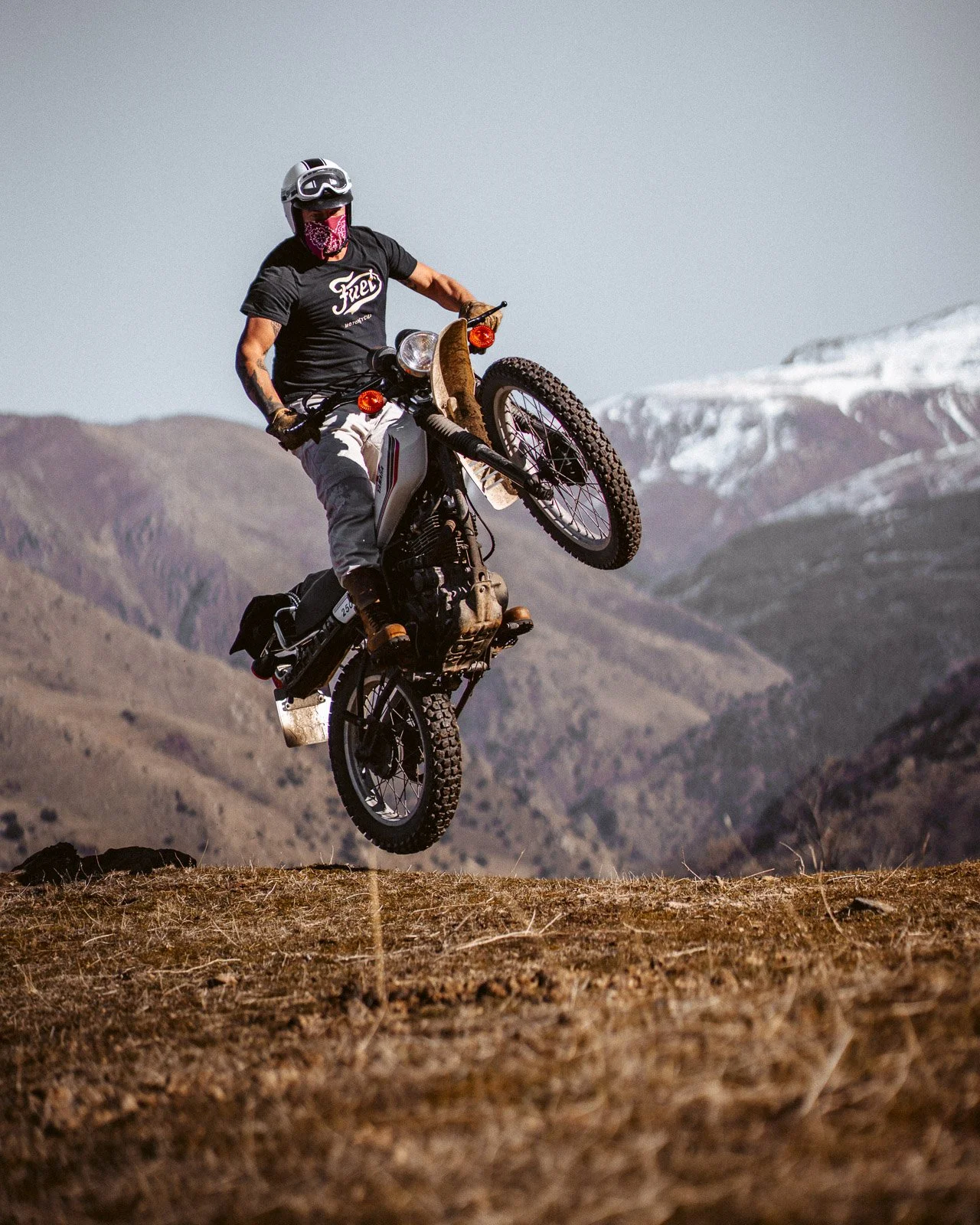 Ein Mann fährt mit einem Motorrad im Offroad-Gelände, hebt das Vorderrad beim Springen, mit Bergen im Hintergrund.