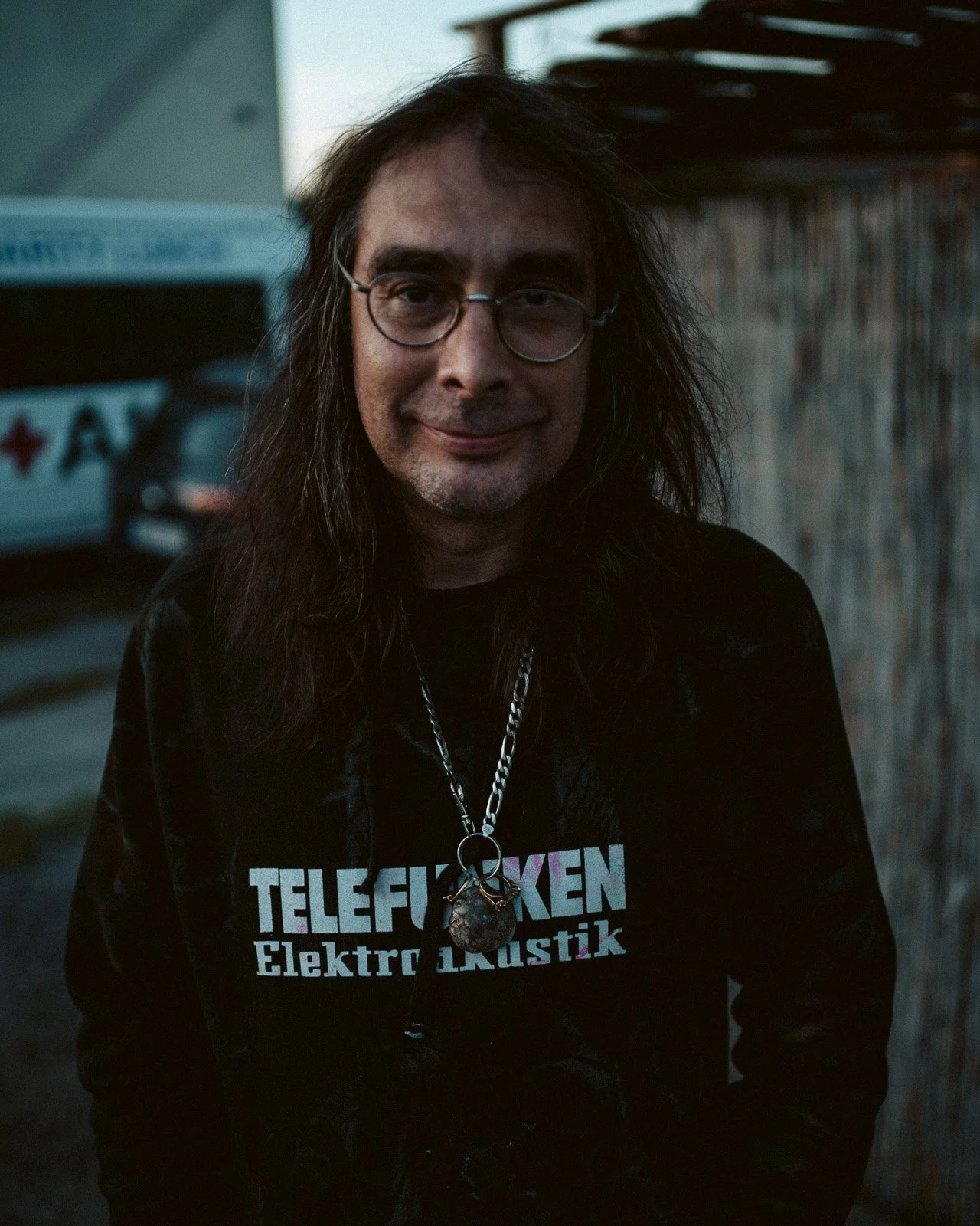 Ein Mann mit langen dunklen Haaren, Brille und einem schwarzen T-Shirt mit der Aufschrift ‚TELEFONEN Elektronik‘ steht im Freien vor einer Holzwand, trägt eine Halskette mit Anhänger.