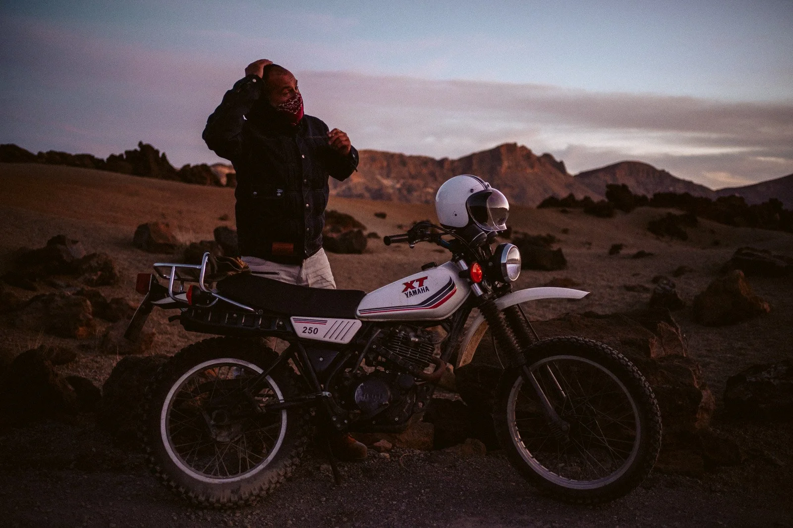 Eine Person steht neben einem weißen Yamaha XR 250 Motorrad in einer Lava-Wandschaft bei sunset, trägt einen schwarzen Mantel und eine rote Gesichtsmaske.