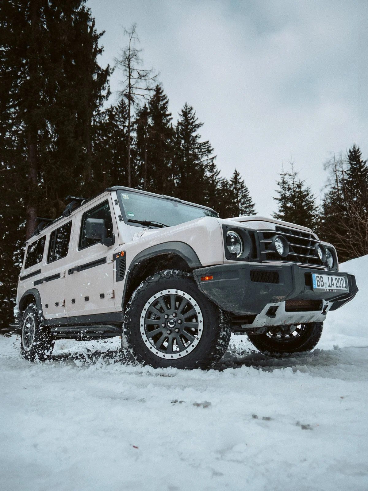 Ein weißer Geländewagen fährt im Schnee, umgeben von Tannenbäumen, bei bewölktem Himmel.