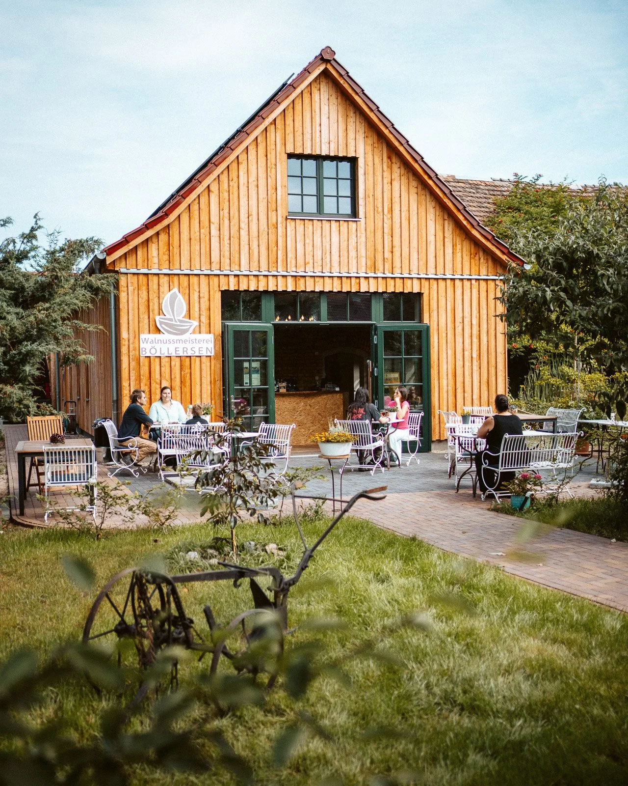 Ein Holzgebäude mit offenen Türen, davor sitzen Menschen auf weißen Stühlen im Garten, einige unter Bäumen, bei einem Kaffee. Es gibt einen historischen Pflug im Vordergrund und eine Beschilderung mit der Aufschrift "Walnussmanufaktur Böllersen".