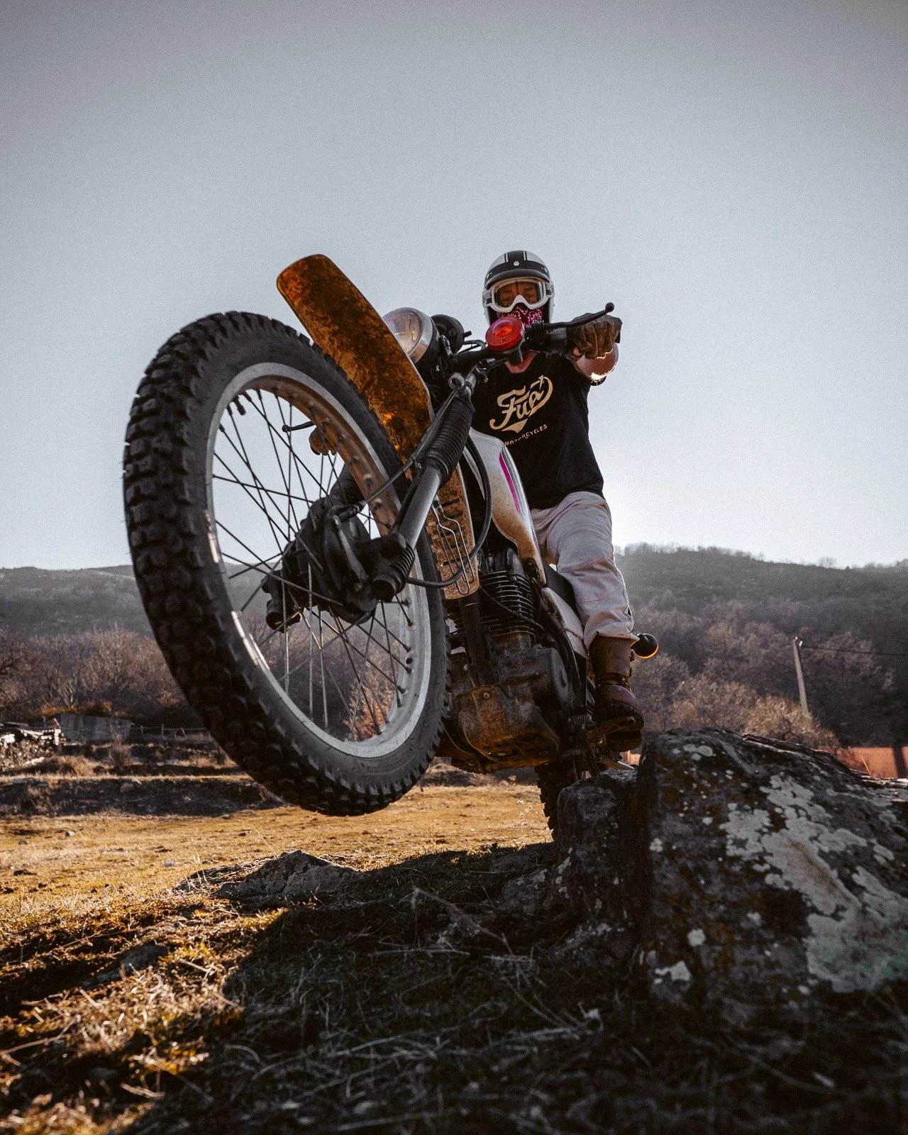Motorradfahrer trägt einen Helm, trägt einen schwarzen T-Shirt mit einem Logo, fährt mit einem alten Motorrad auf einem unebenem Gelände und hebt die Vorderrad hochüber einem Felsen