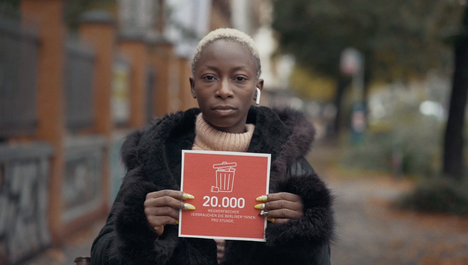 Eine Frau mit kurzen, hellen Haaren hält ein rotes Plakat mit einem weißen Mülleimer und der Zahl 20.000, sowie deutschem Text, stehend auf einer Straße, umgeben von Bäumen und Gebäuden.