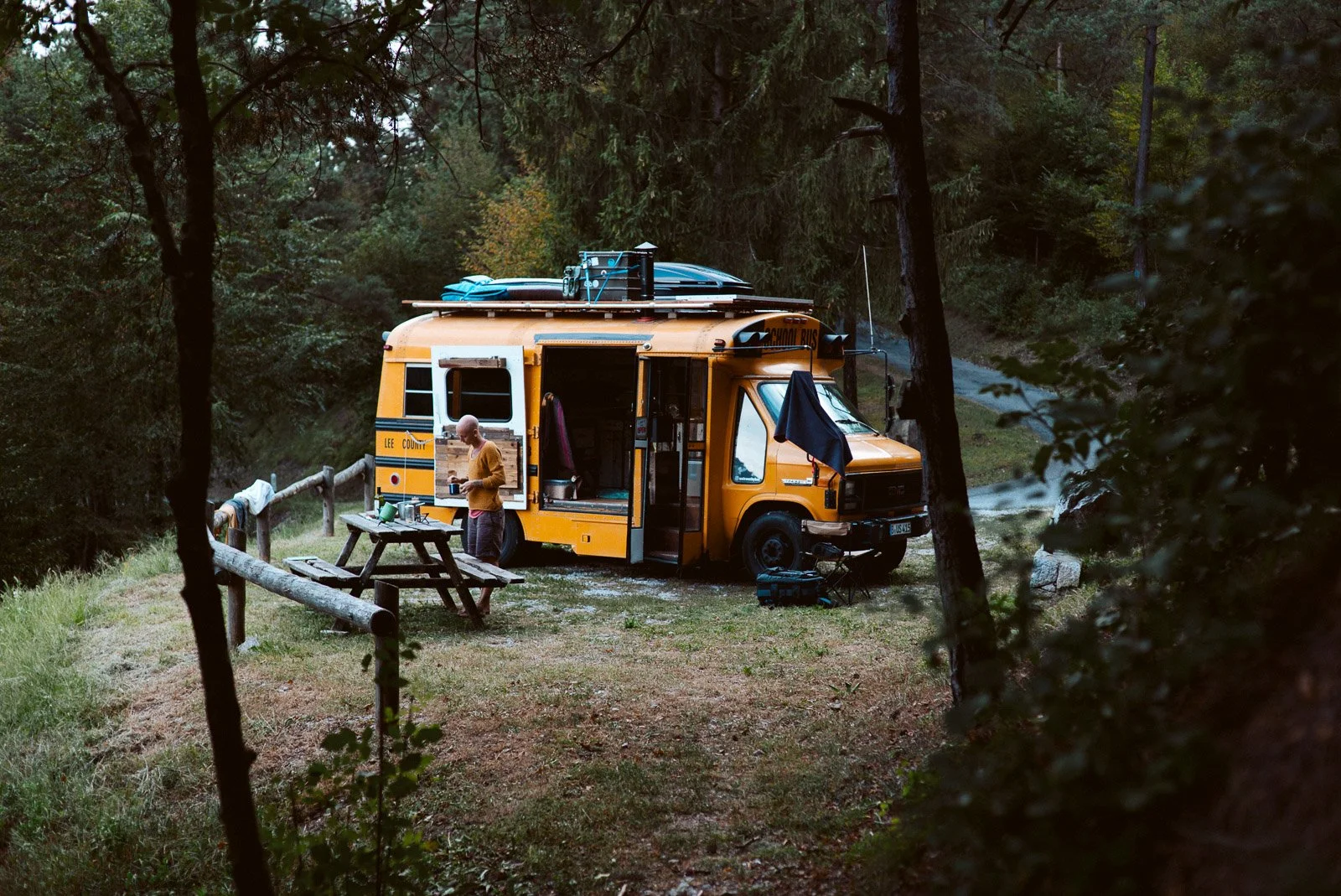 Eine Person steht vor einem gelben Schulbus, der in einer Waldumgebung geparkt ist. Es wird im Freien gearbeitet, mit einem Tisch und verschiedenen Gegenständen daneben.